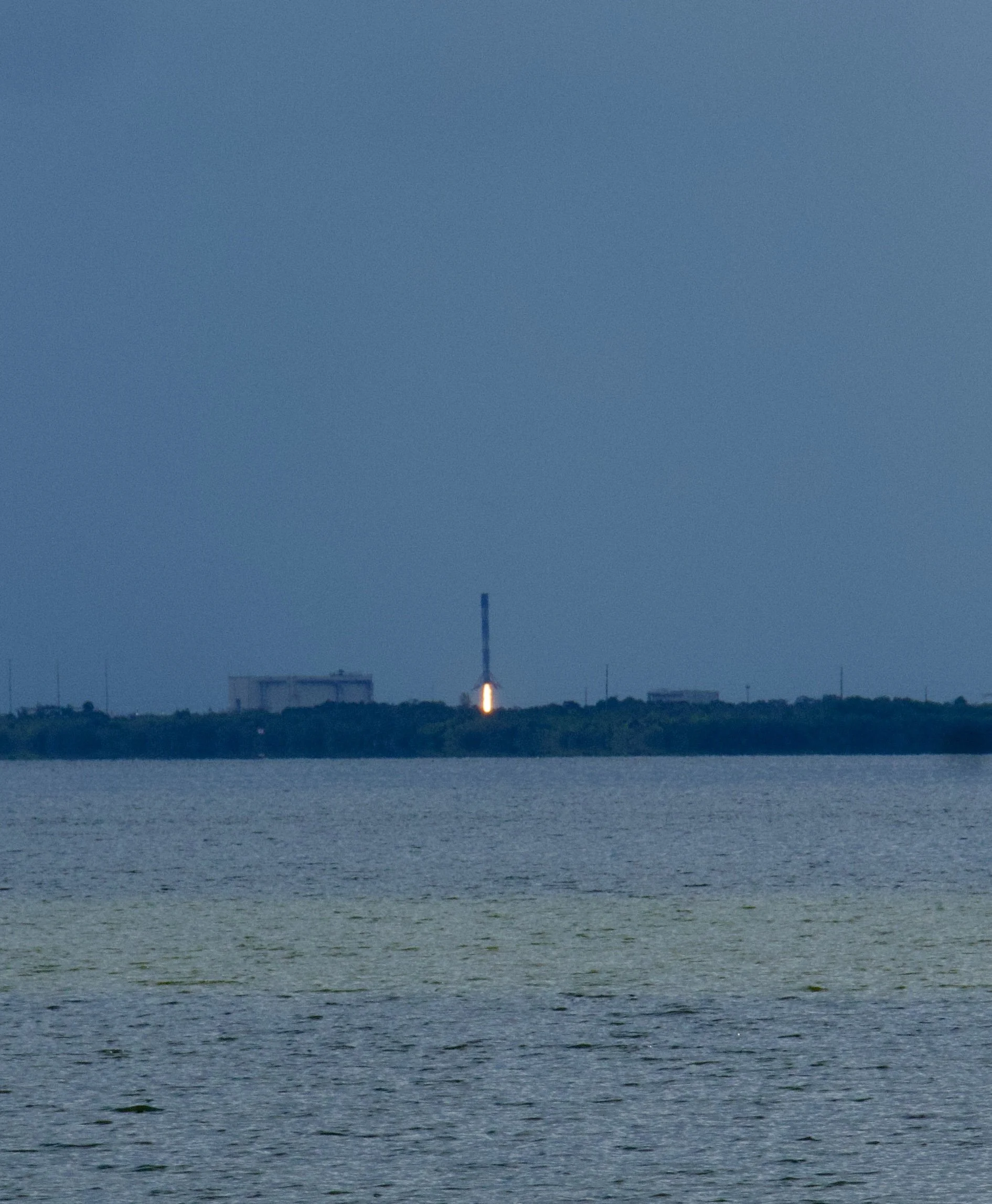 Rocket launching from a launchpad near a body of water, with a blue sky in the background.