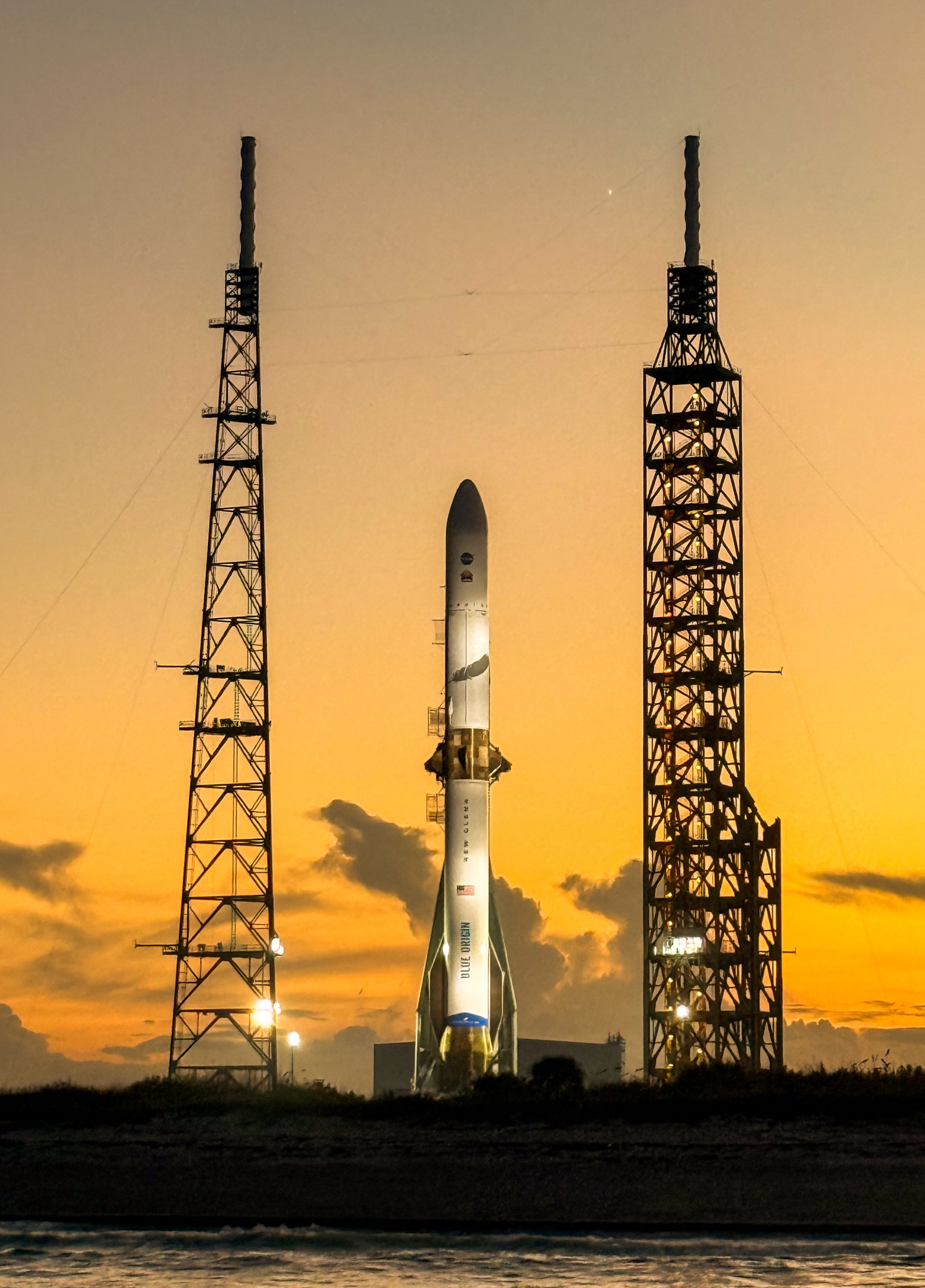 A SpaceX Blue Origin rocket standing on a launch pad between two tall towers during sunset.
