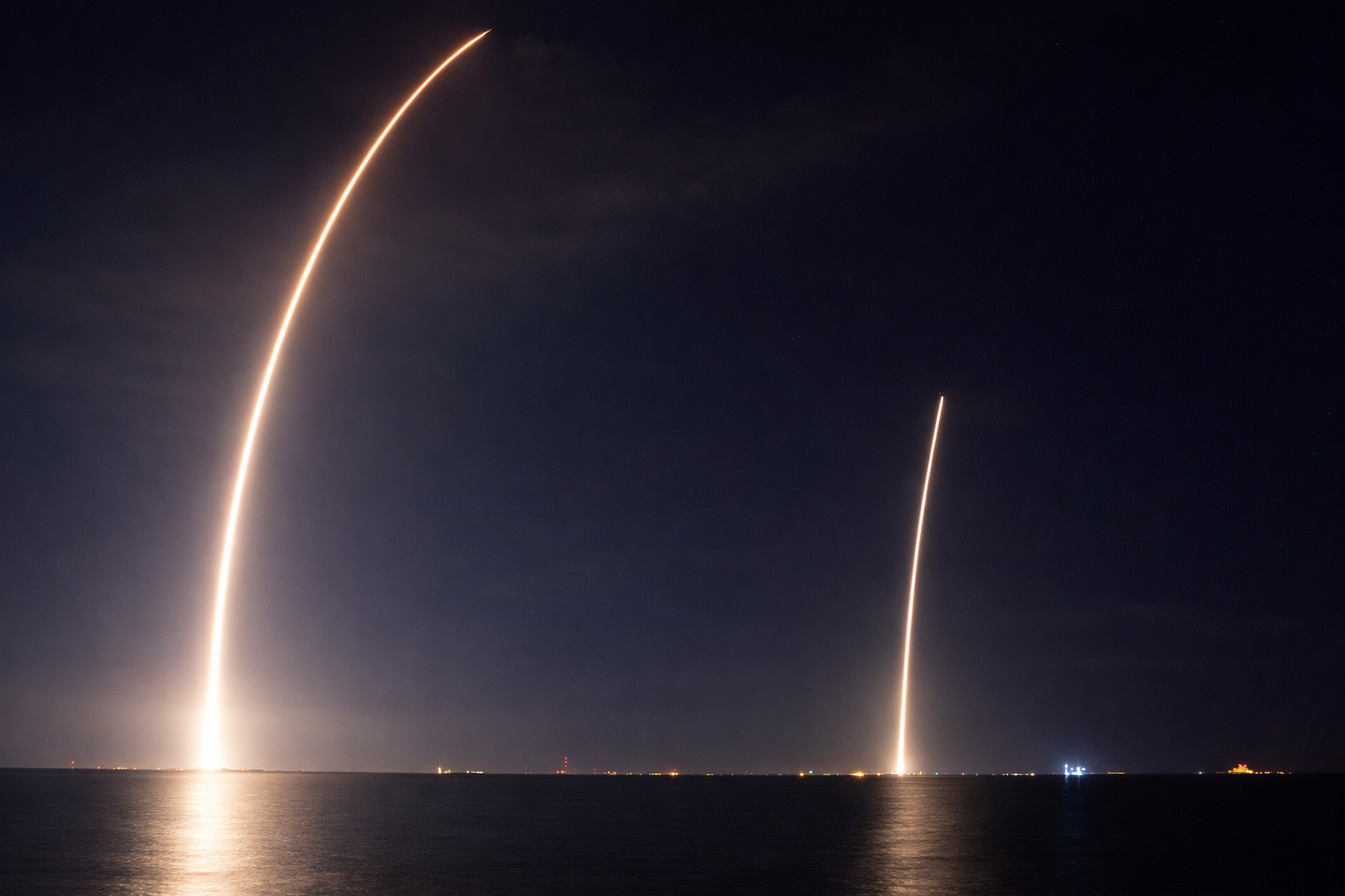 Two rockets launching into the night sky over a body of water, leaving bright trails.
