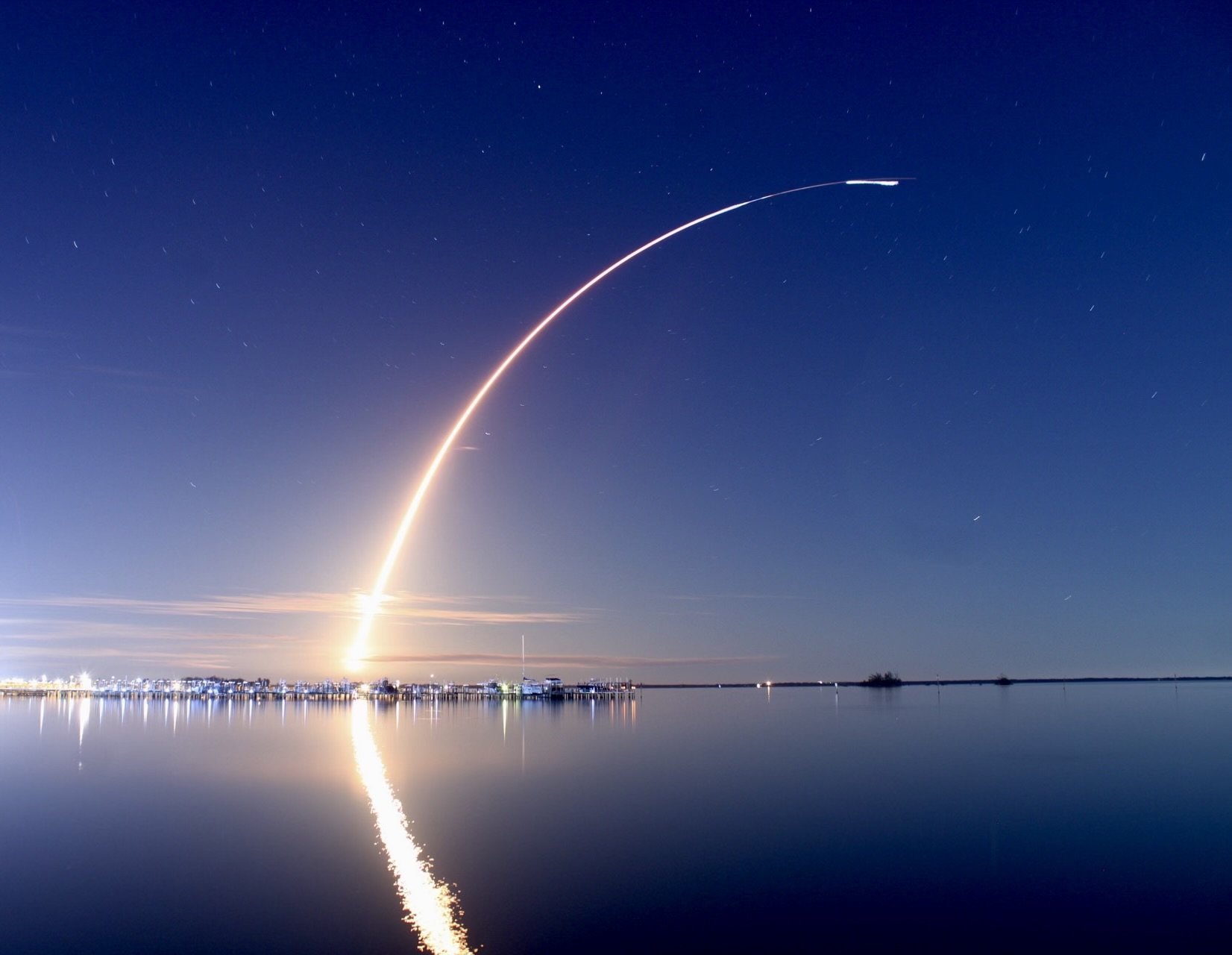 Night sky over a body of water with a rocket launching, leaving a bright trail and reflection on the water