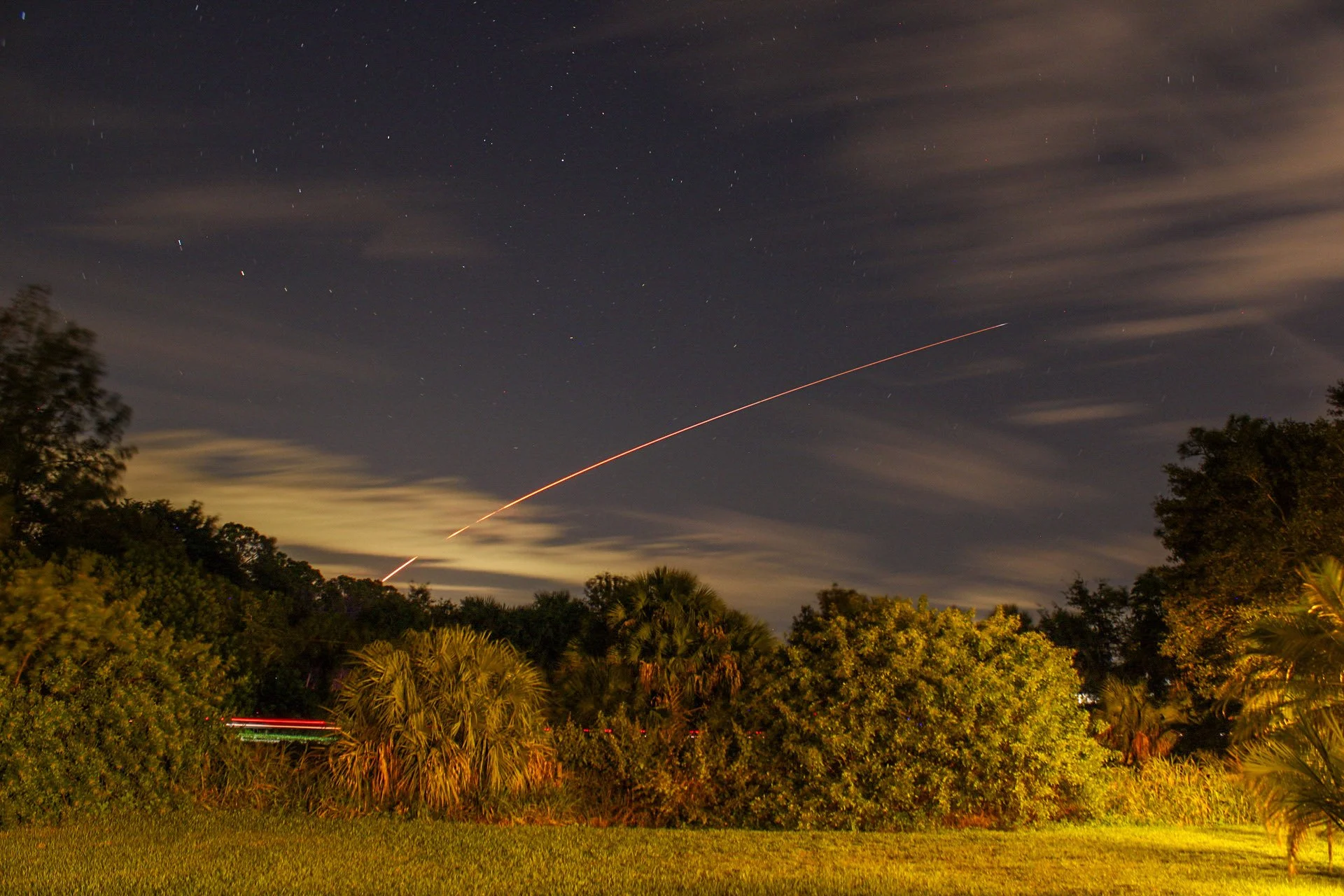 Nighttime scene of a forest with trees and bushes, with a streak of light across the sky indicating a launching rocket or missile, and stars visible in the cloudy sky.