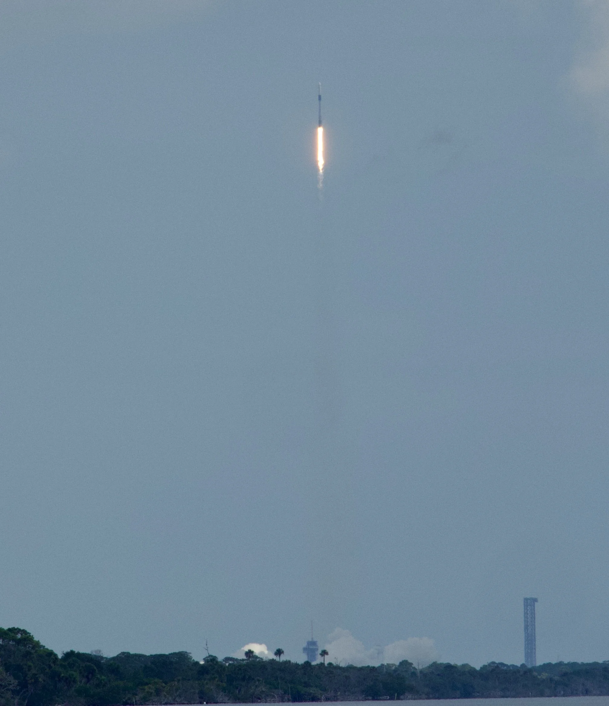 A rocket is launching into the sky, leaving a bright trail of fire and smoke. The launch site is visible at the bottom with trees and structures, including a tower, against a cloudy sky.