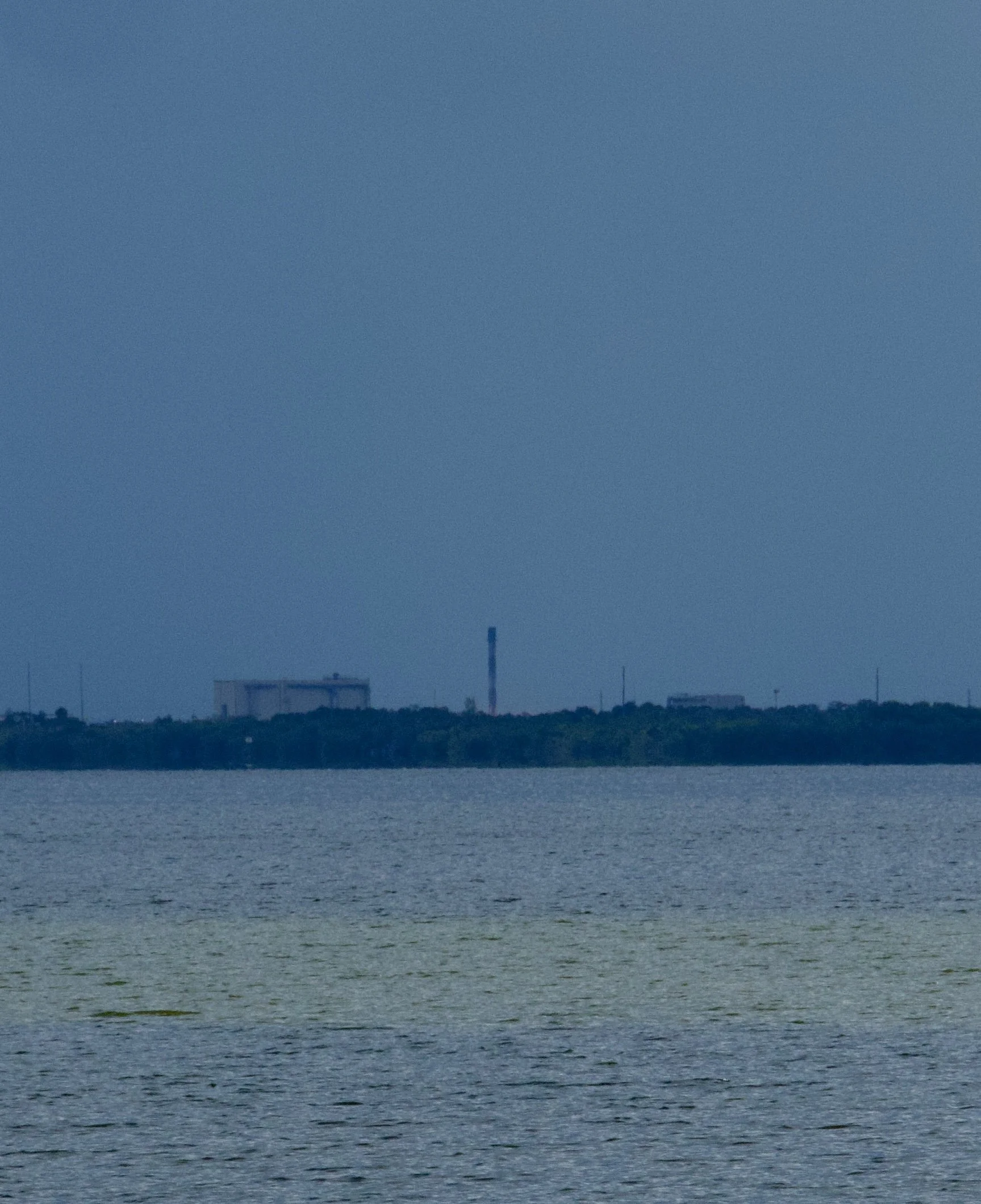 A body of water with a distant shoreline featuring industrial buildings and a tall smokestack under a blue sky.