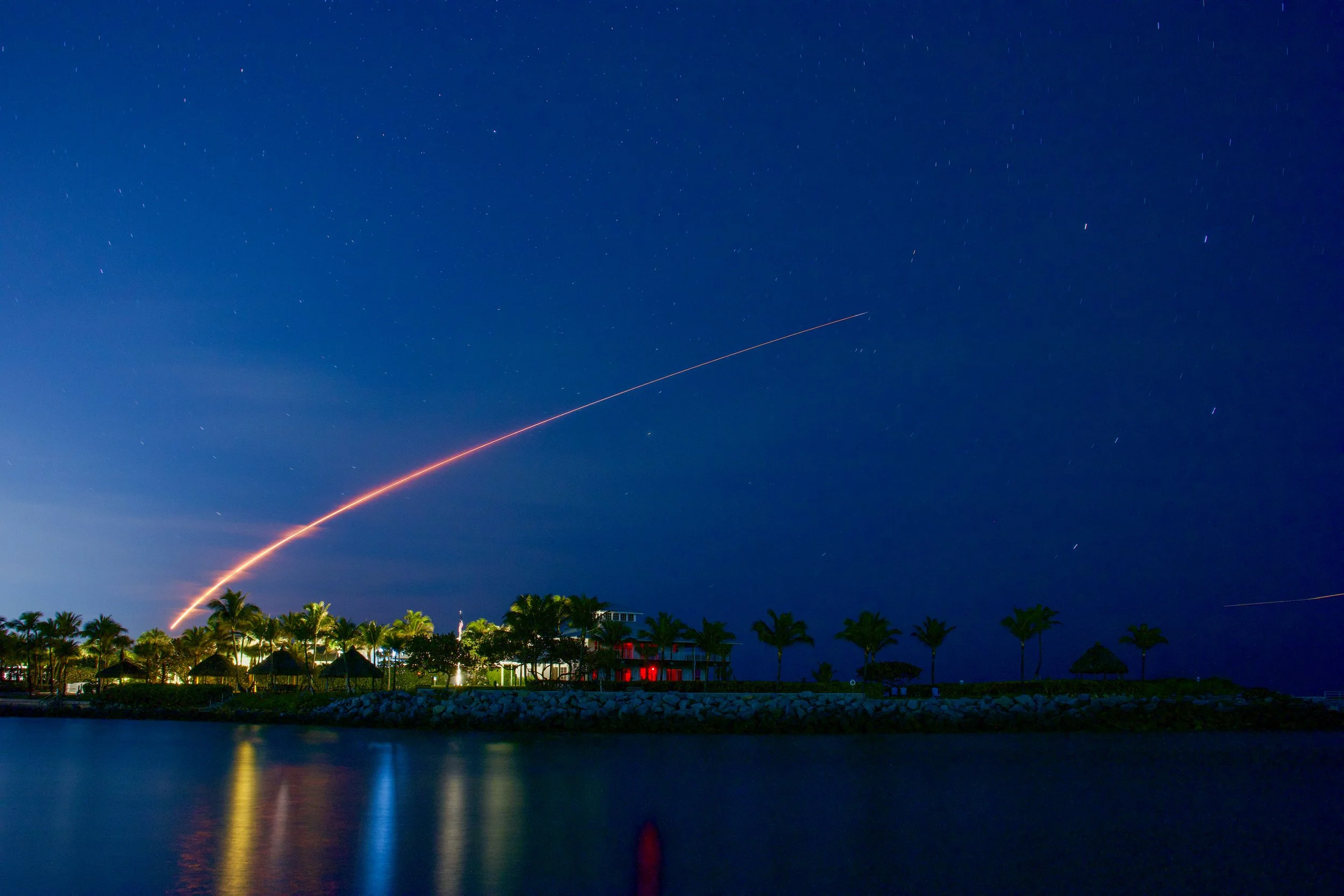 Nighttime scene of a fireworks display over a tropical resort with palm trees, thatched-roof umbrellas, and calm water reflections.