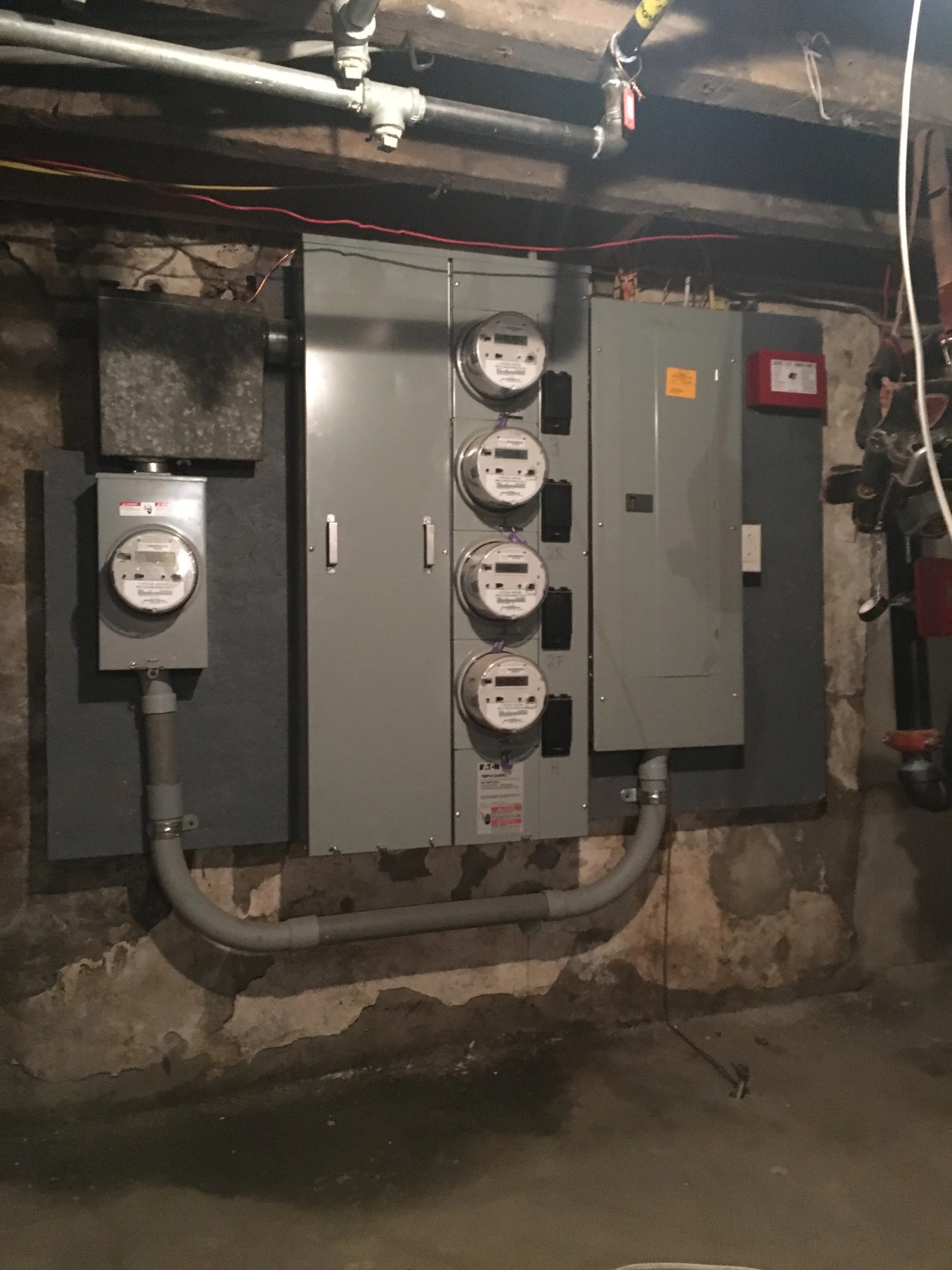 Electrical meters and panels on a basement wall with exposed pipes and conduit.