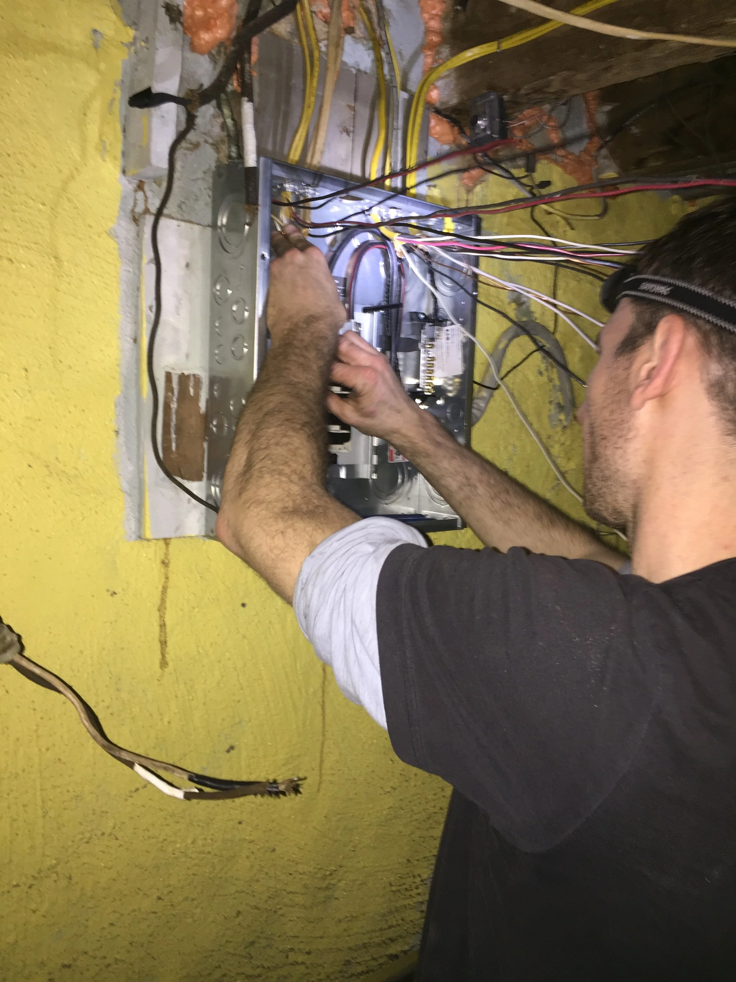 A person working on wiring inside an electrical panel mounted on a yellow-painted wall.