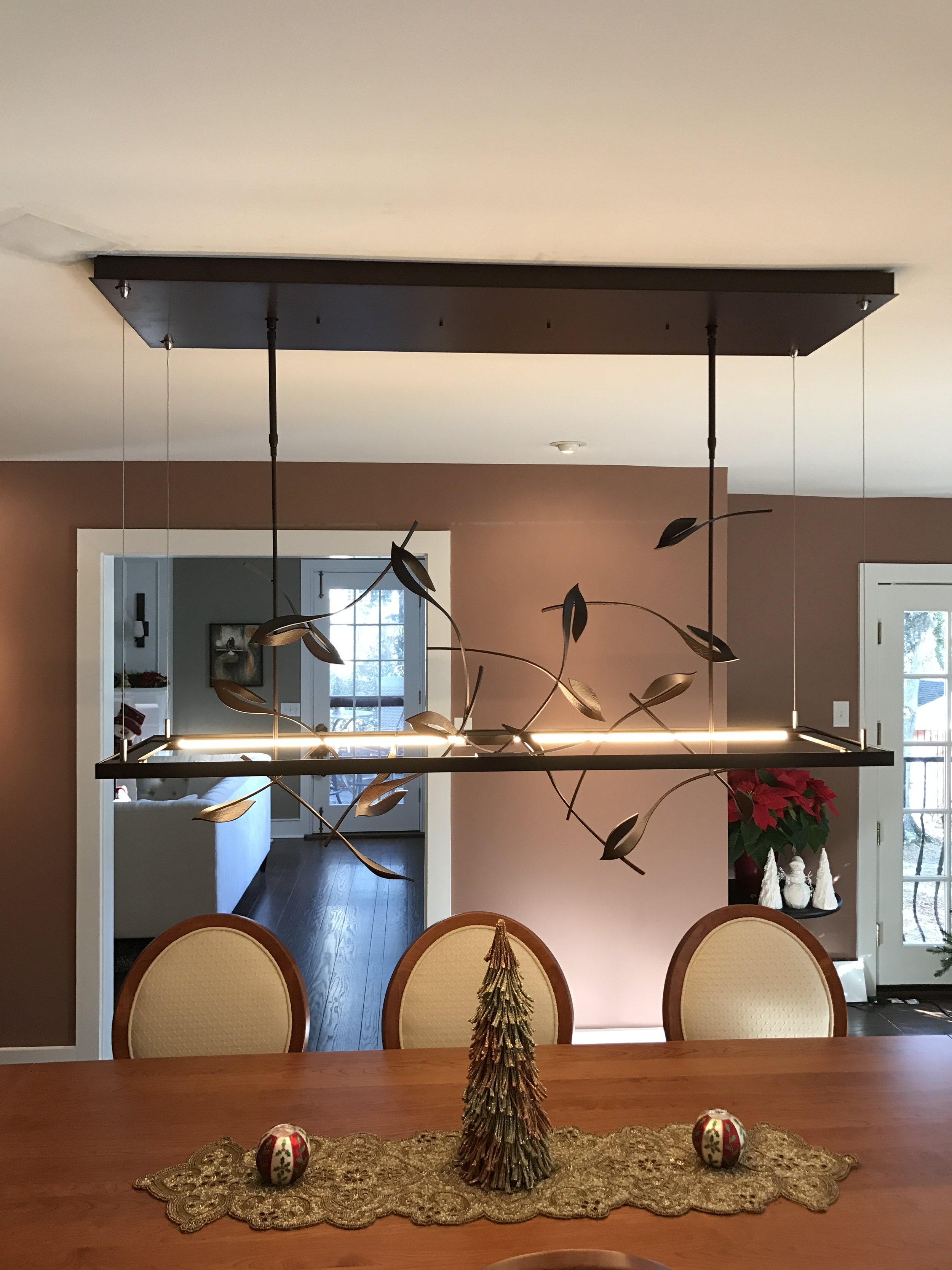A dining room table with a decorative gold table runner and Christmas ornaments, including a small gold star tree and two decorated eggs, in front of four beige upholstered chairs. A chandelier with leaf-shaped lights hangs above, and a poinsettia is in the corner near a door with glass panels.