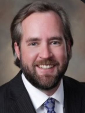 A smiling man with brown hair and a beard, wearing a dark suit, white shirt, and a patterned tie.