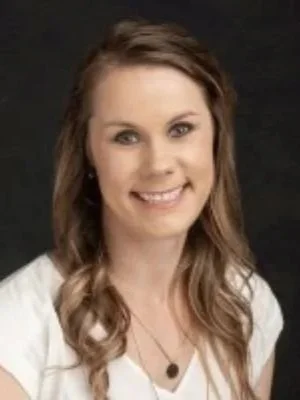 A young woman with long wavy brown hair smiling at the camera, wearing a white top and a necklace, against a dark background.