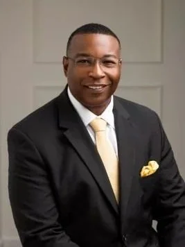 A man in a black suit with a light-colored tie and pocket square, smiling and standing indoors.