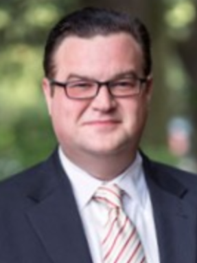 A man in a suit and tie with glasses, outdoors with a green background.