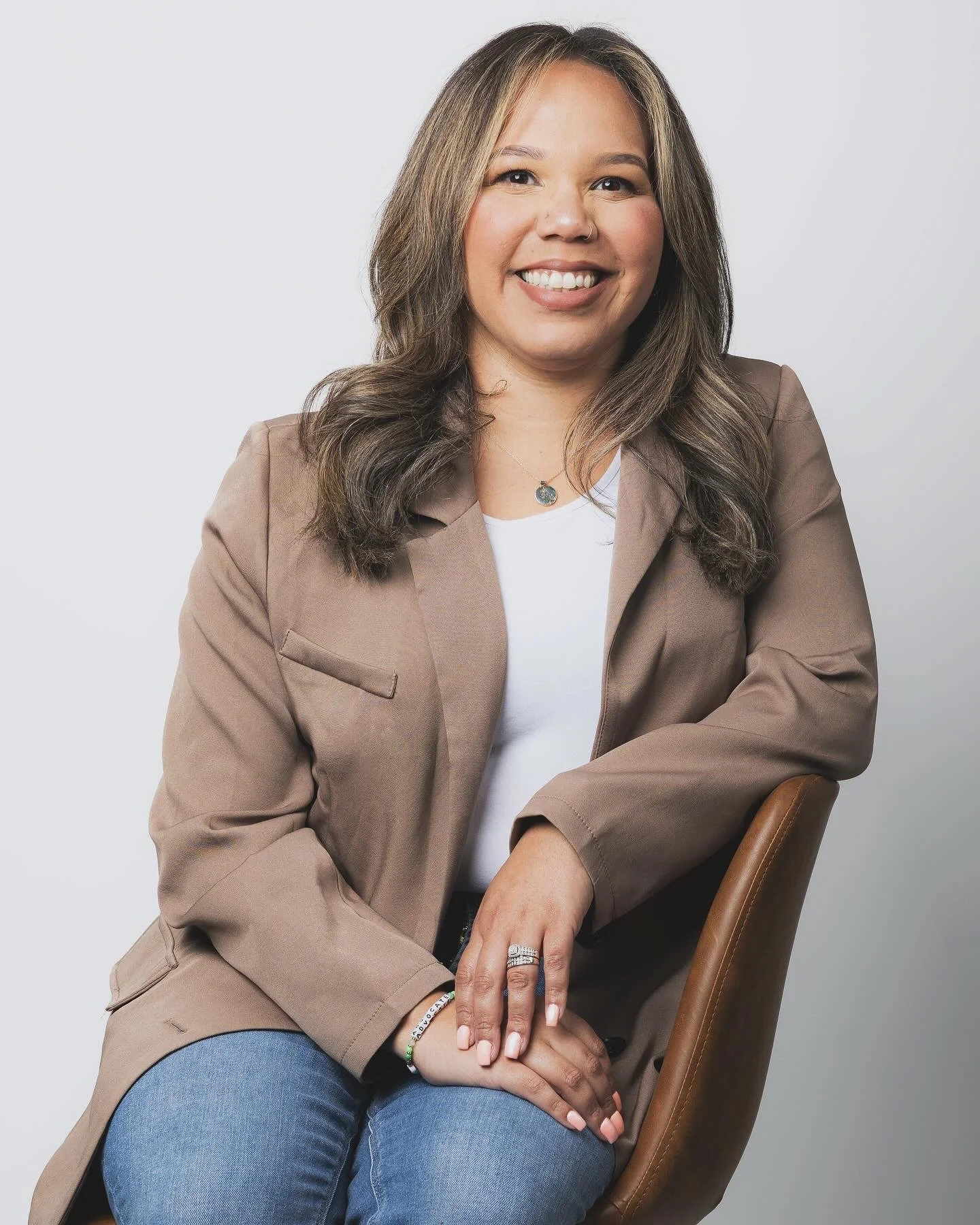 A woman with shoulder-length brown hair sitting on a brown chair, smiling, wearing a tan blazer, white top, and blue jeans.