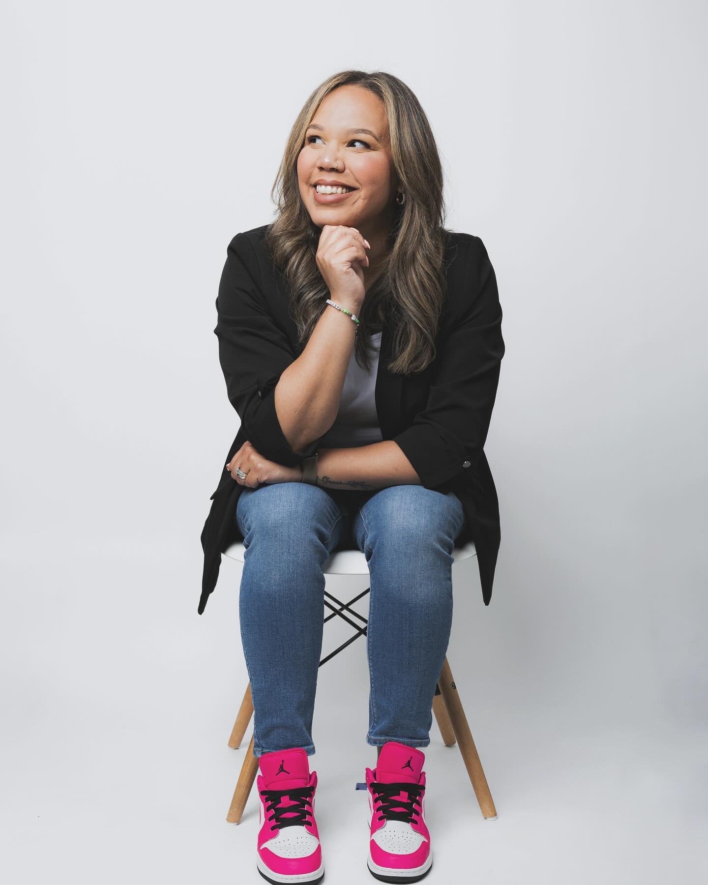 Woman with long wavy hair sitting on a white chair, smiling, wearing a black blazer, white shirt, blue jeans, and pink sneakers, against a plain light gray background.