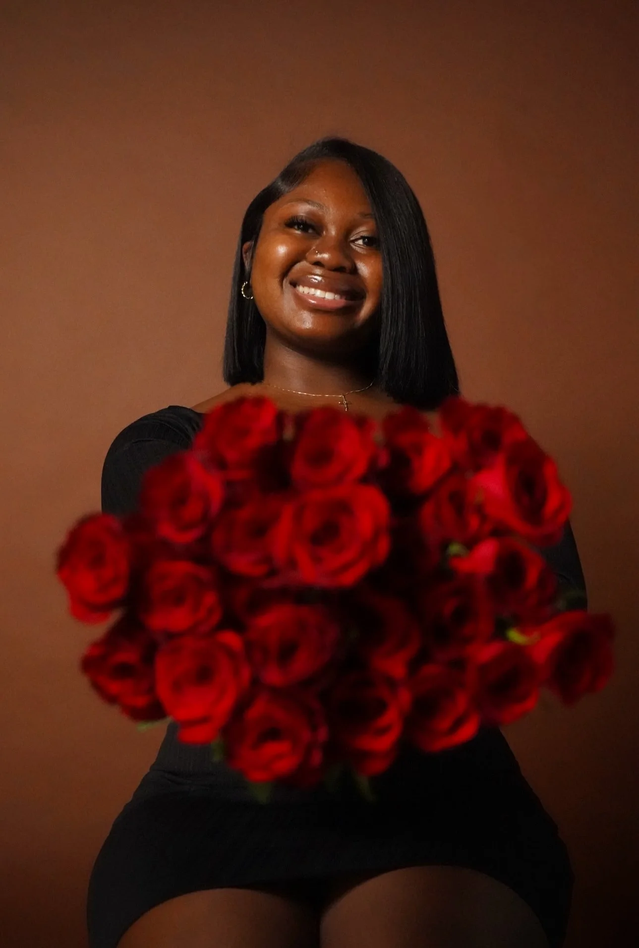 A smiling person holding a large bouquet of red roses against a brown background.