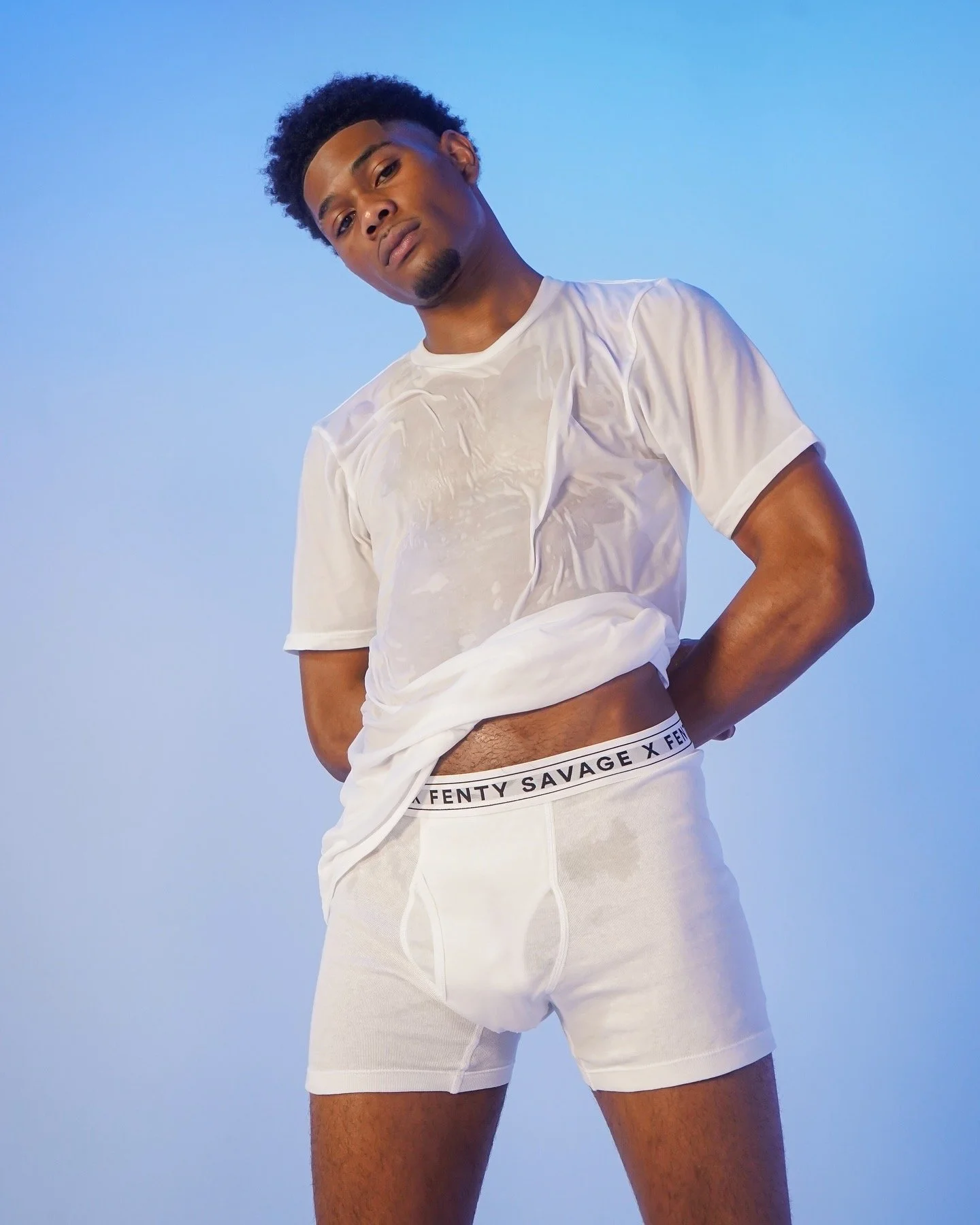 Model posing in white Savage X Fenty underwear and sheer white shirt against blue background.