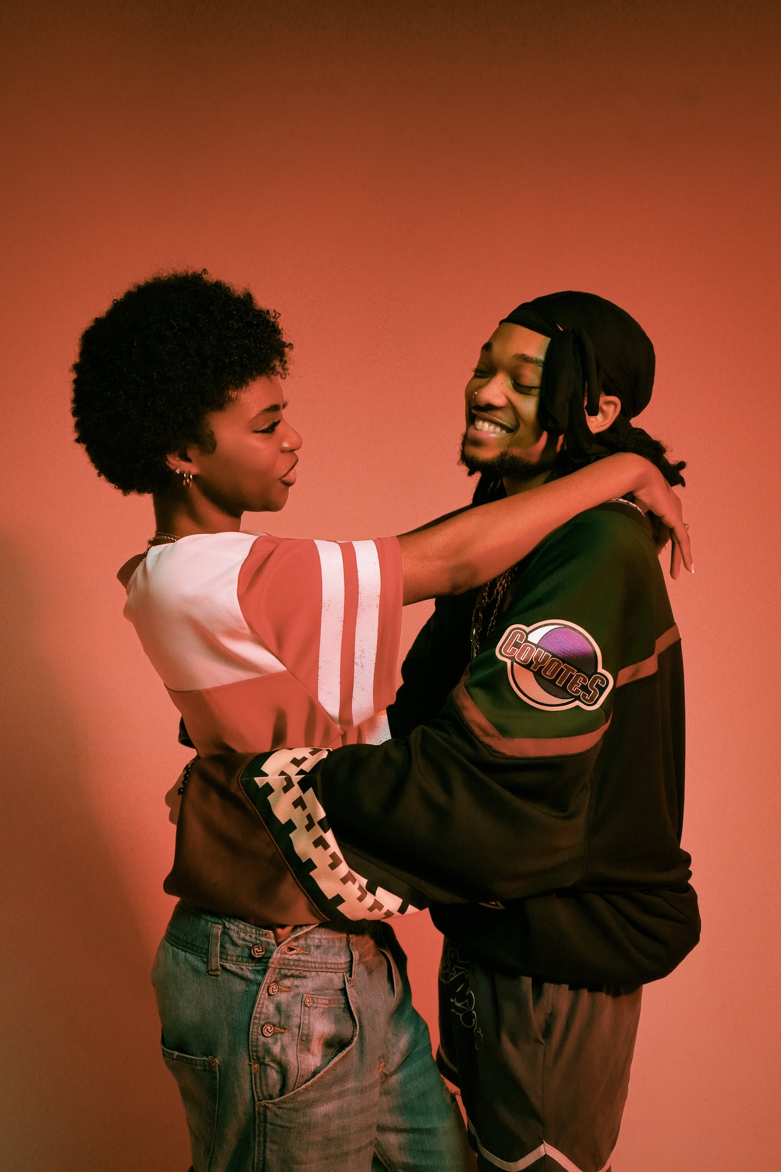 A couple embracing with smiles, against a warm-colored background. The person on the left is wearing a striped shirt, and the person on the right is in a jersey with "Coyotes" on the sleeve.
