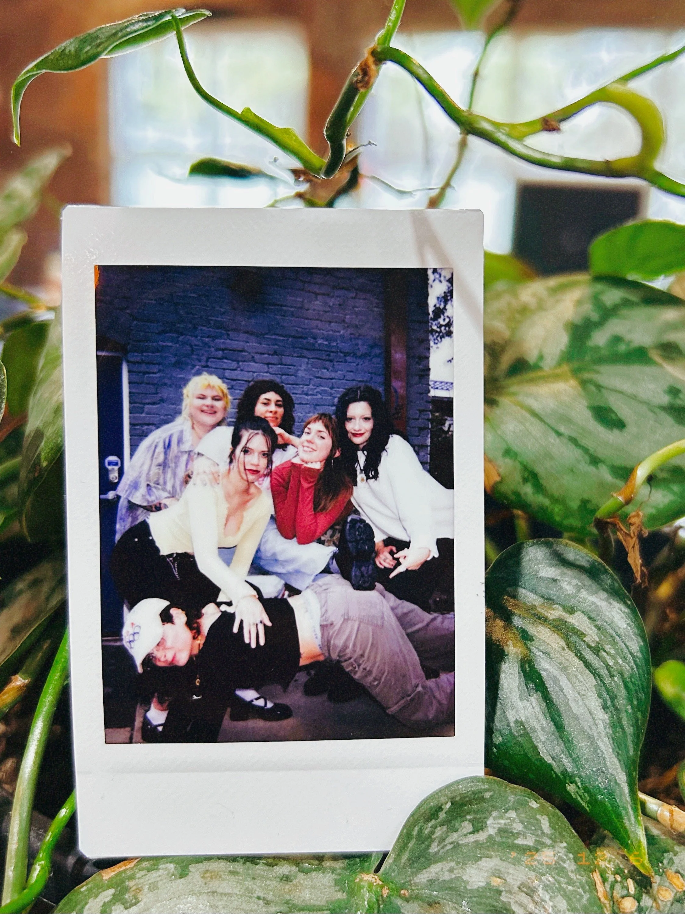 A group of six young women posing together indoors against a dark brick wall background. One woman is lying on the floor with her head resting on another woman's lap, while the others are gathered around, smiling and making playful gestures.