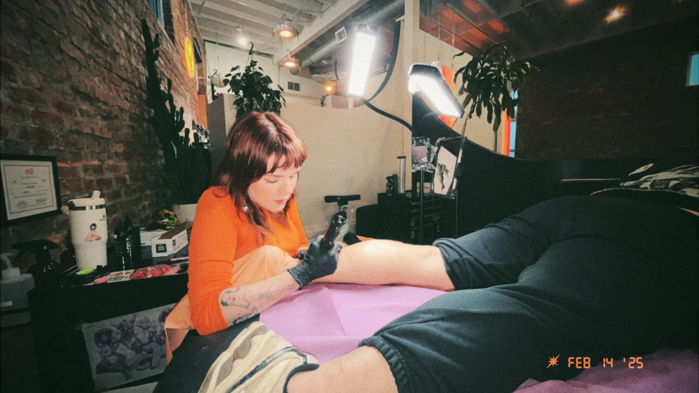 Tattoo artist with shoulder-length red hair and black gloves ink a tattoo on a client's arm in a tattoo studio with brick walls, plants, and art on display.