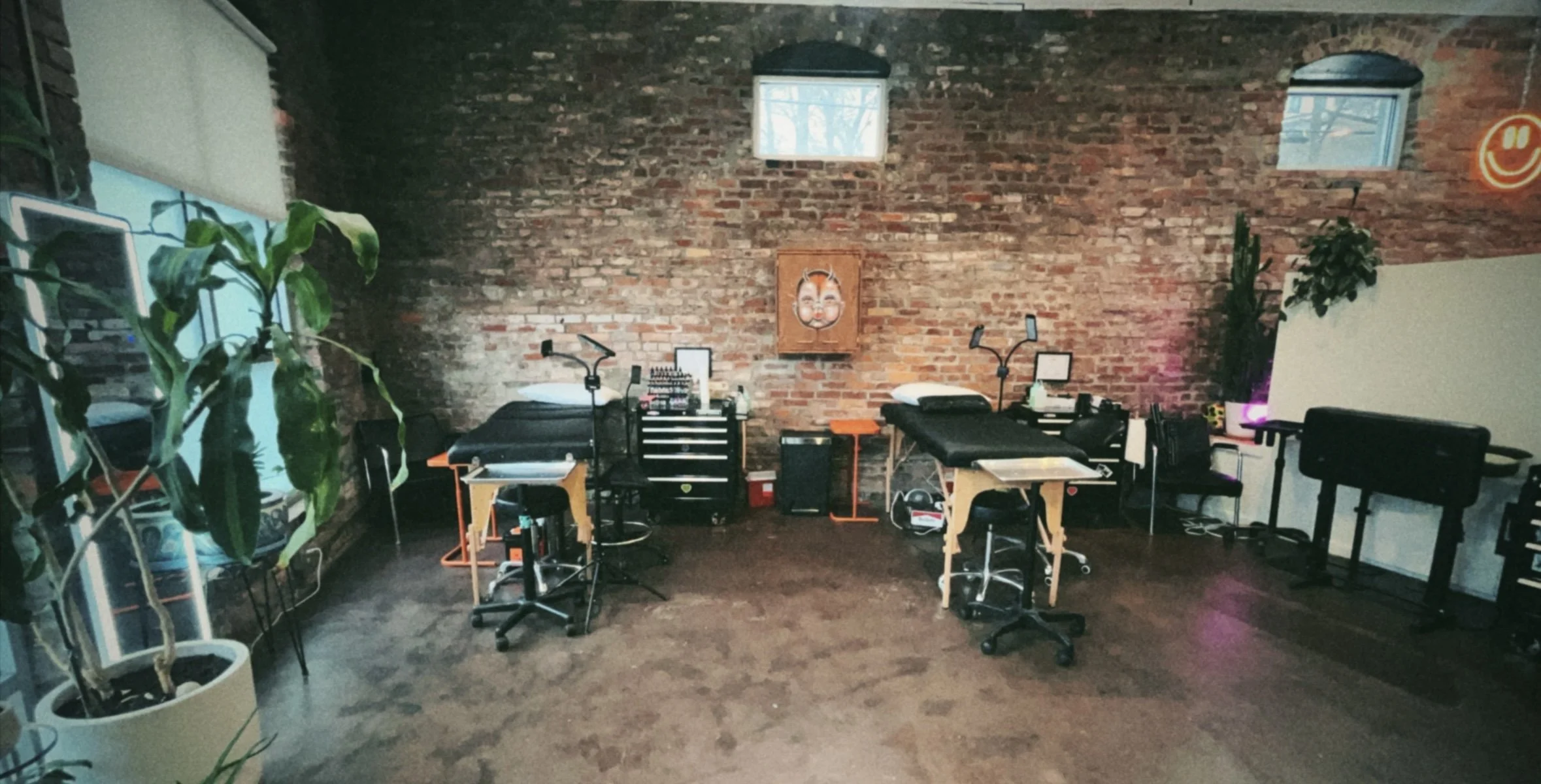A tattoo studio with three tattoo chairs, work tables, and various tattoo equipment against an exposed brick wall. There are two small windows near the ceiling, potted plants, and a neon smiley face sign.