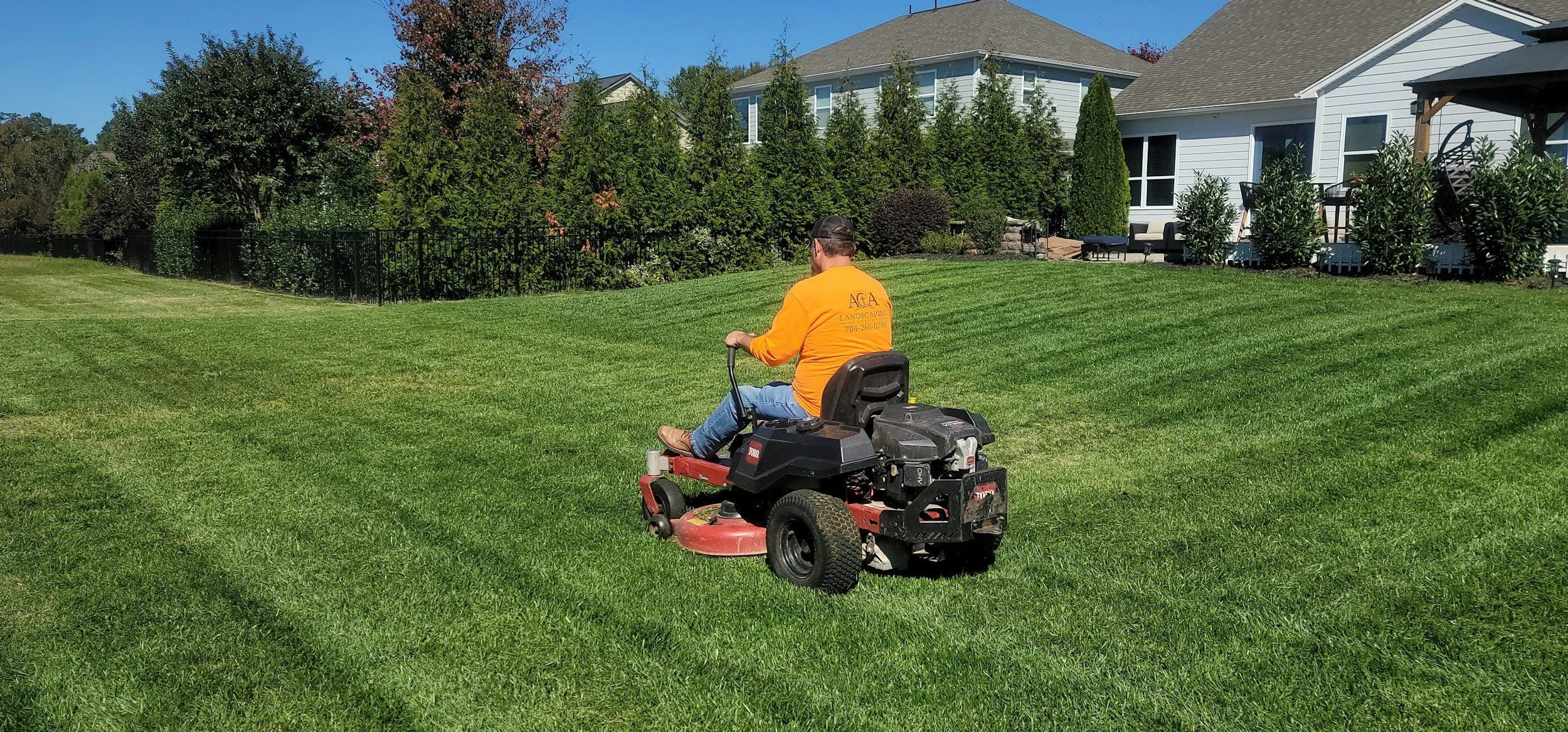 A man operating a riding lawn mower on a well-manicured backyard lawn with houses and trees in the background.