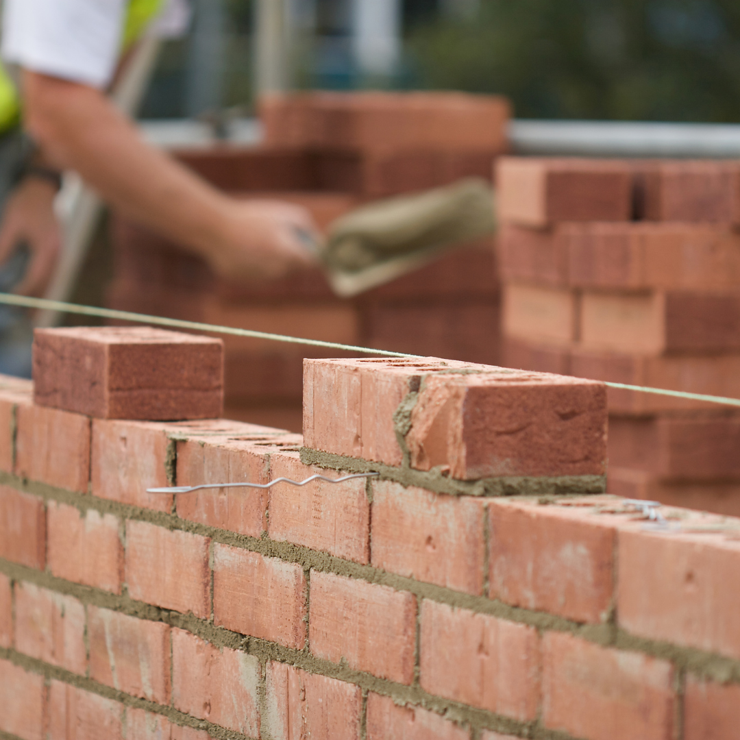Bricklayer using a trowel to lay bricks on a wall construction site with mortar, with stacks of bricks in the background.