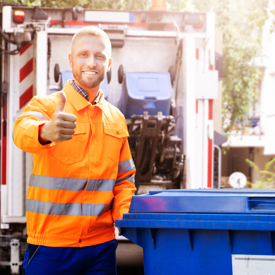 Smiling worker in an orange safety jacket giving a thumbs-up next to a blue recycling bin and a garbage truck