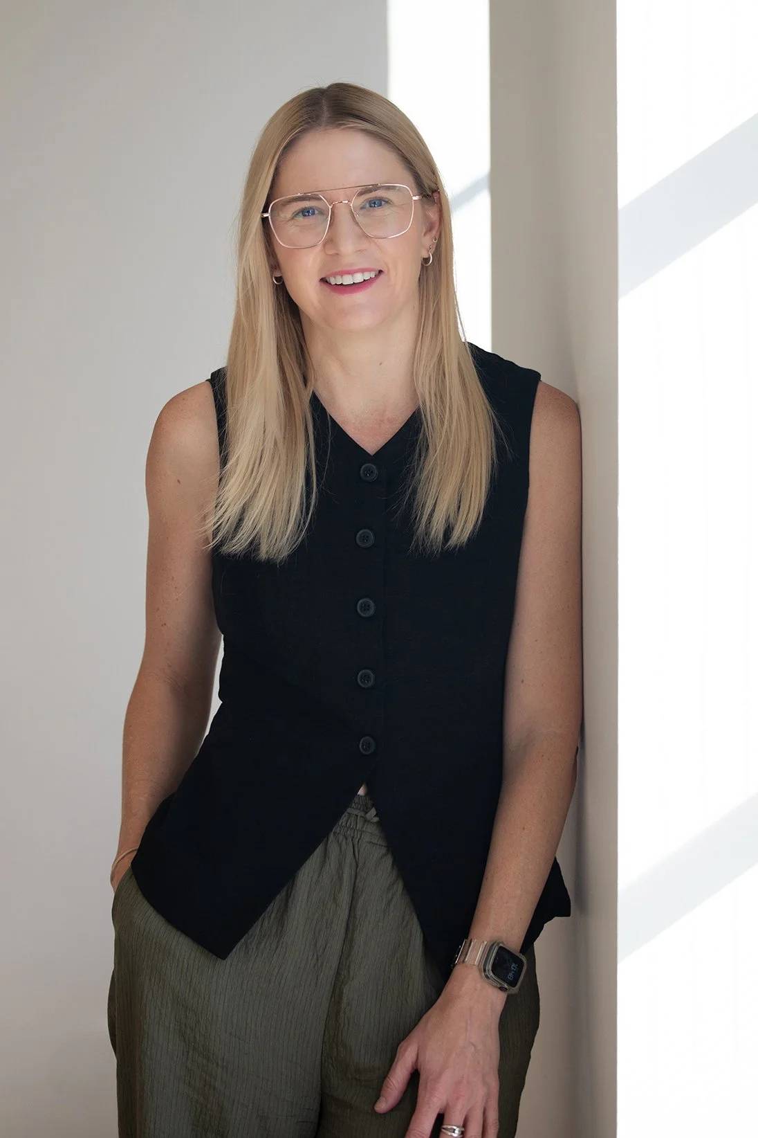 Woman with shoulder-length light brown hair and glasses, wearing a sleeveless white top and black pants, leaning against a white wall with natural light.