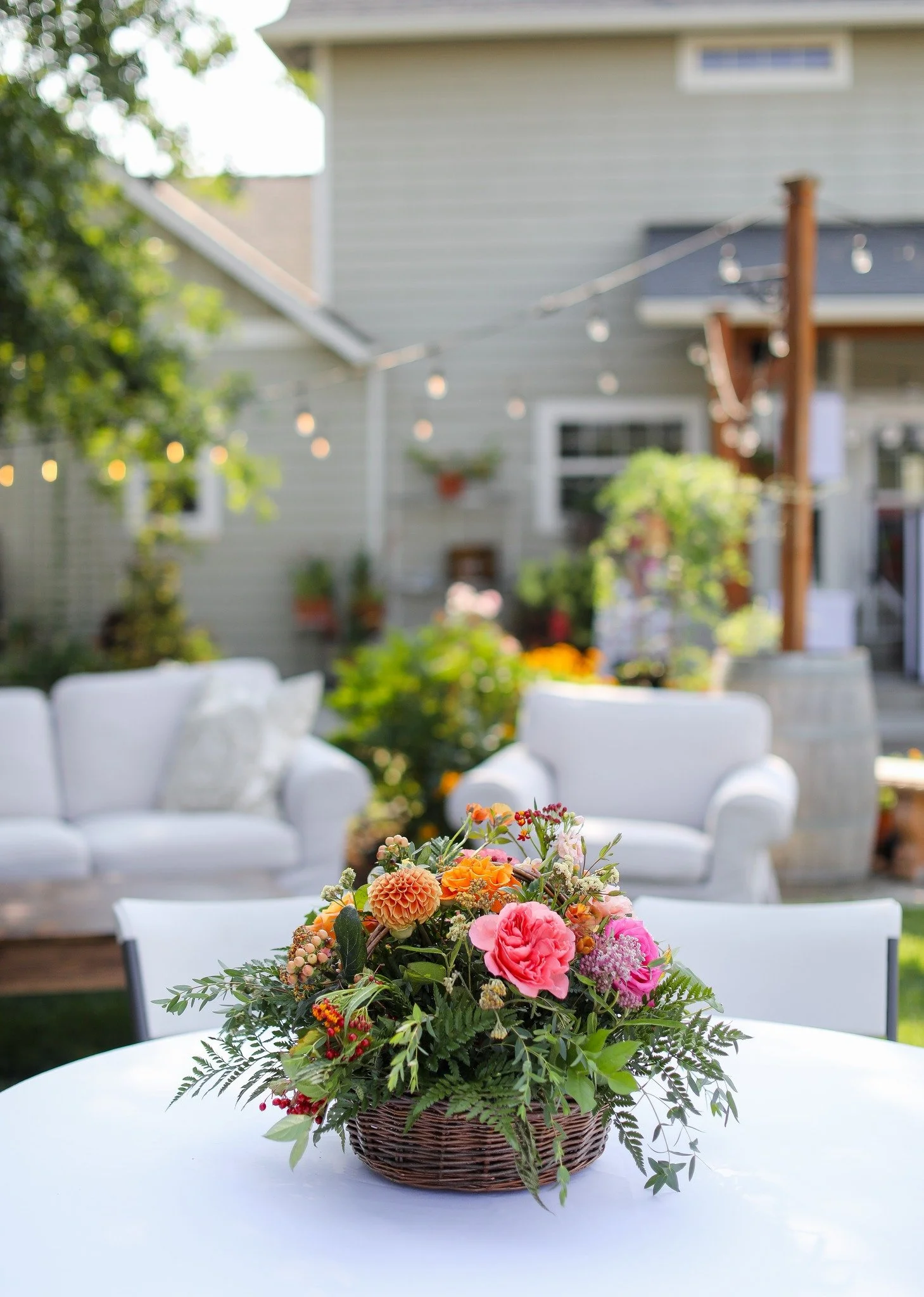 A wicker basket filled with a colorful flower arrangement on a white outdoor table, with blurred white outdoor furniture and a house with string lights in the background.