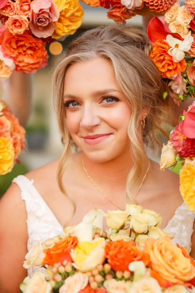 A woman with blonde hair and blue eyes smiling at the camera, surrounded by a vibrant flower arch and holding a bouquet of mixed orange, yellow, and cream-colored roses.