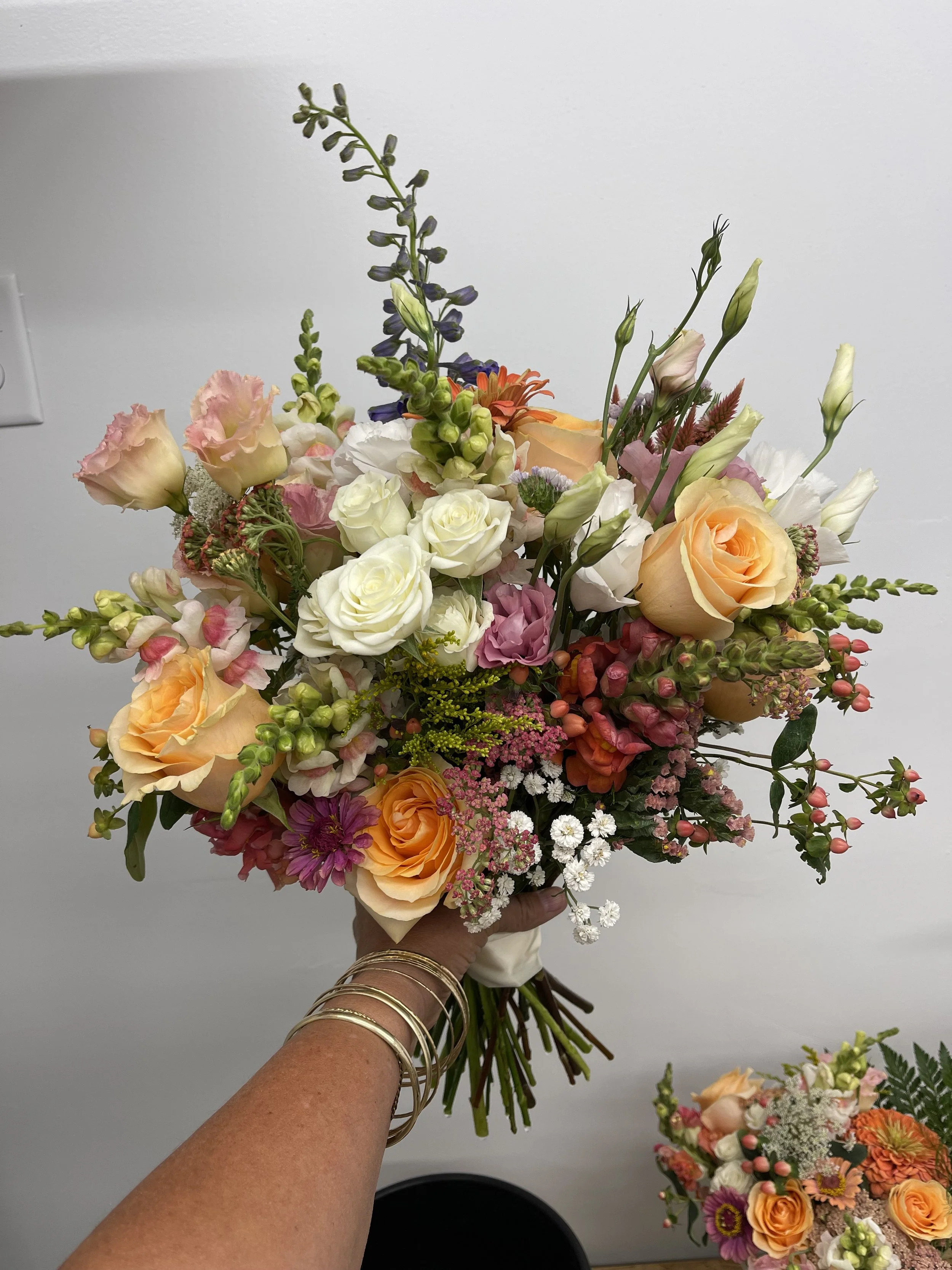 Hand holding a colorful mixed flower bouquet with roses, snapdragons, and various wildflowers against a plain background.