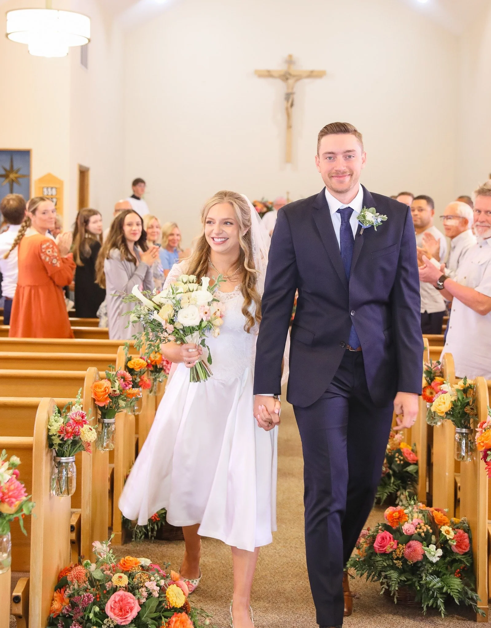 A newlywed couple walking down the aisle after their wedding ceremony inside a church. The bride is holding a bouquet of flowers and both are smiling. Guests are clapping and smiling in the background.