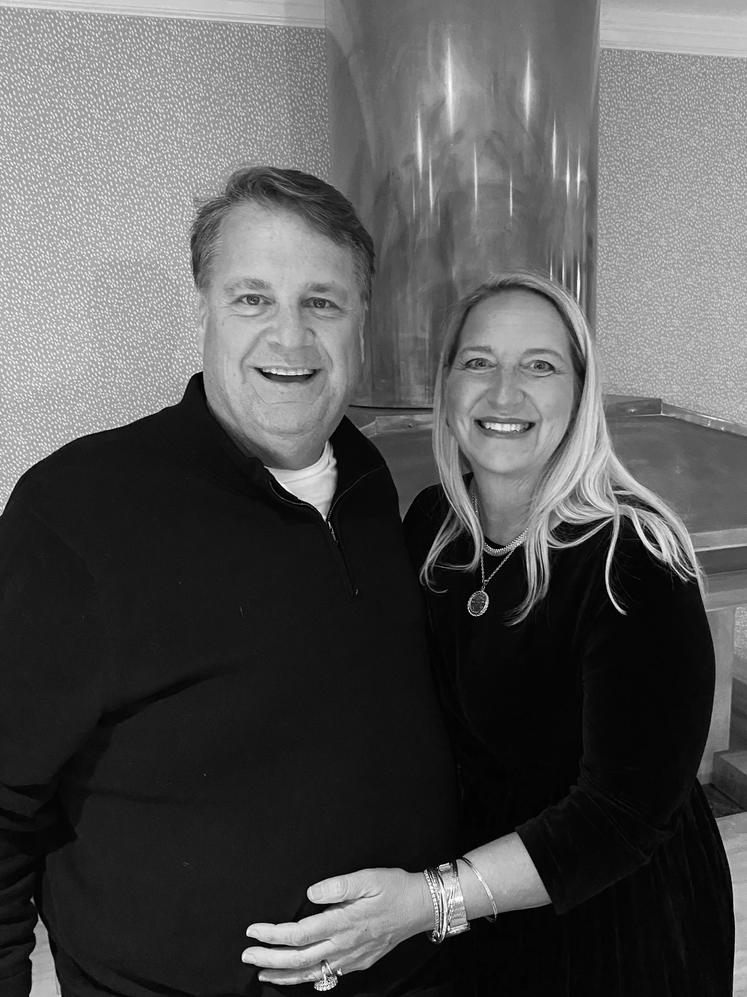 A black and white photo of a smiling man and woman standing closely together, with the woman's hand resting on the man's stomach.