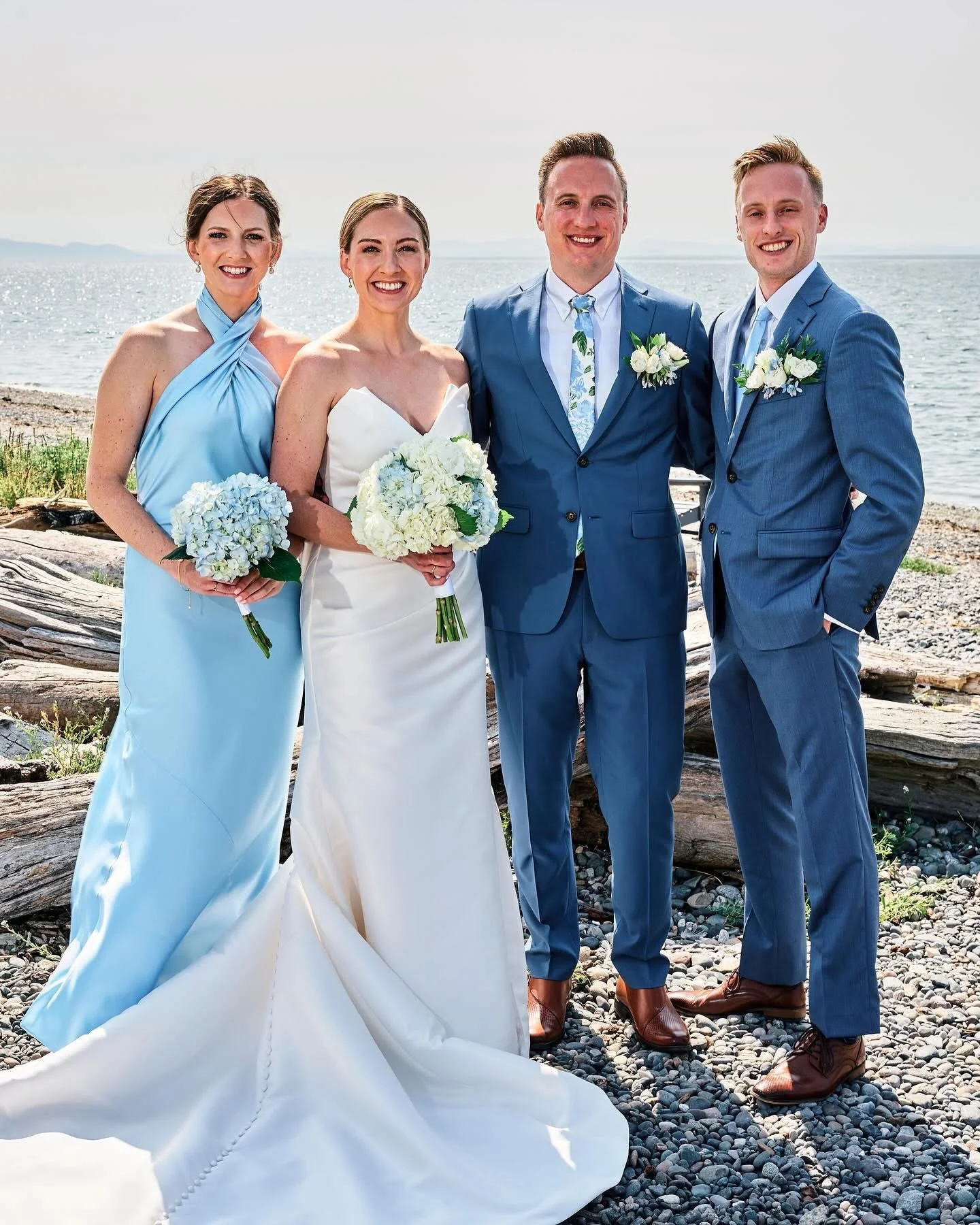 A group of four people in wedding attire standing on a rocky beach with the ocean in the background, smiling at the camera.