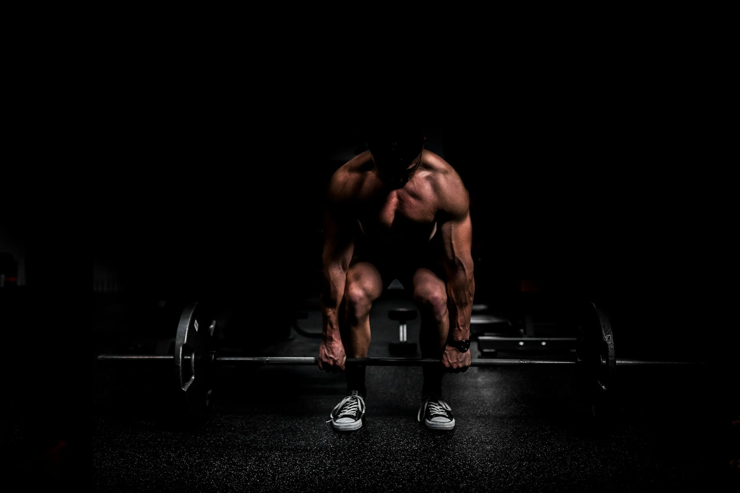 A muscular man in a dark gym setting lifting a barbell with weights on the floor.