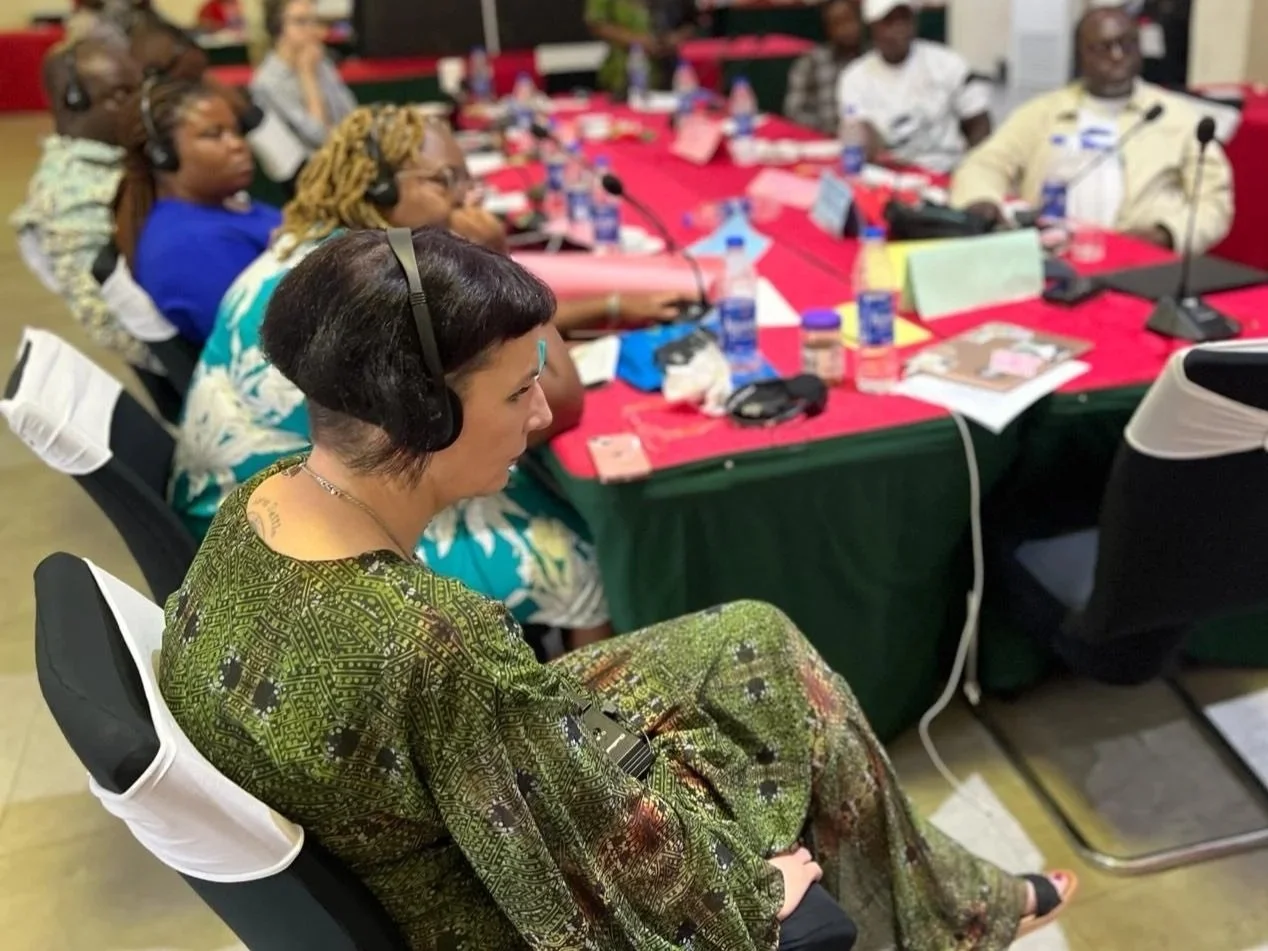 A woman with short dark hair and a necklace, wearing a green patterned dress, sits at a conference table with other attendees. The table has various documents, water bottles, and microphones, and the woman is wearing a headset.