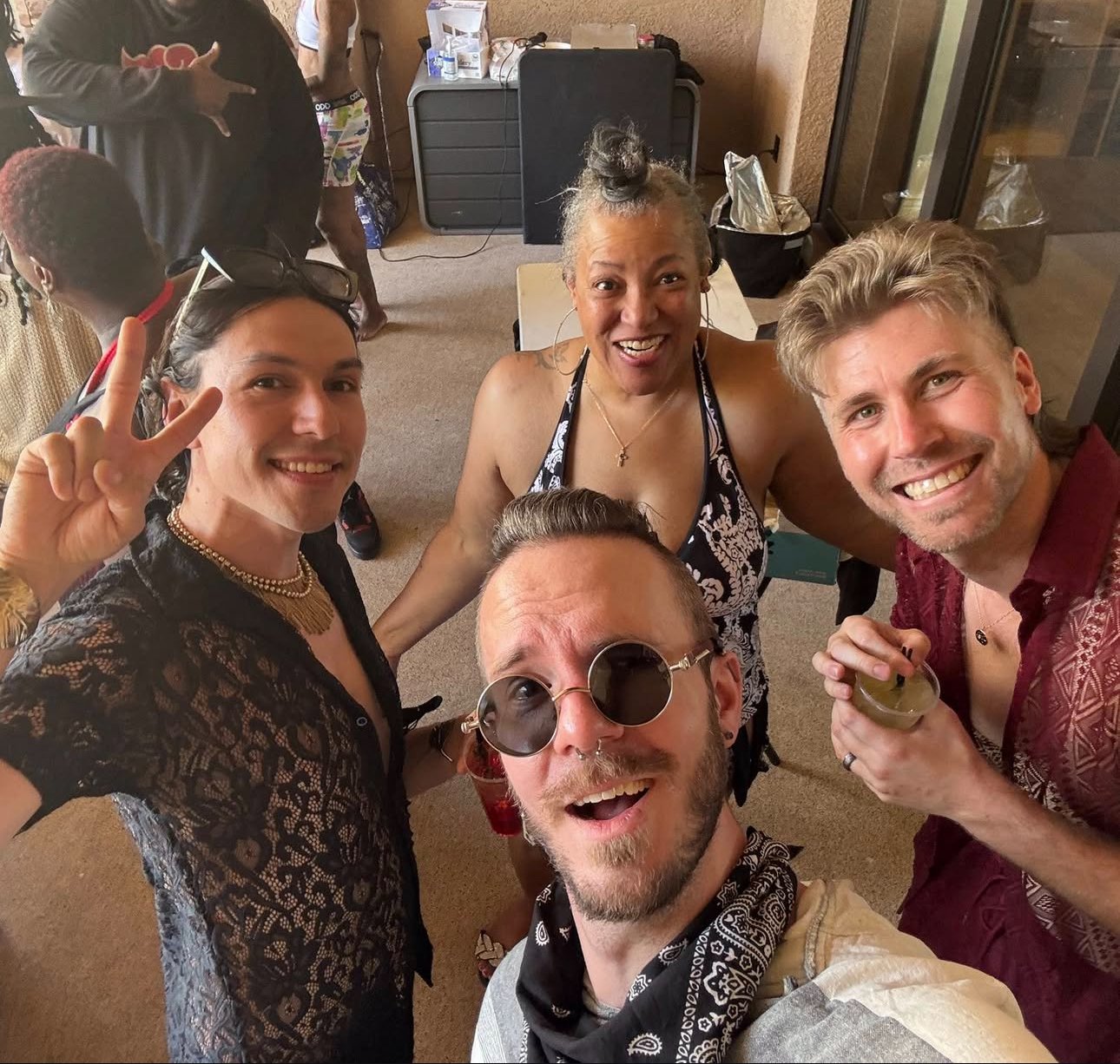 Group of five friends taking a selfie at a social gathering, smiling and showing peace signs. Three men and two women, in a room with a table and other people in the background.