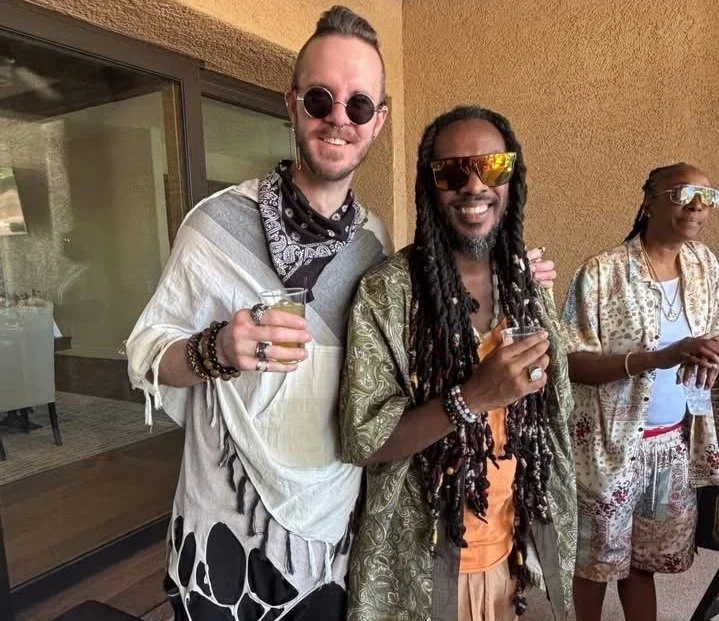 Three people standing outdoors, smiling and holding drinks, wearing sunglasses and casual festival-style clothing.