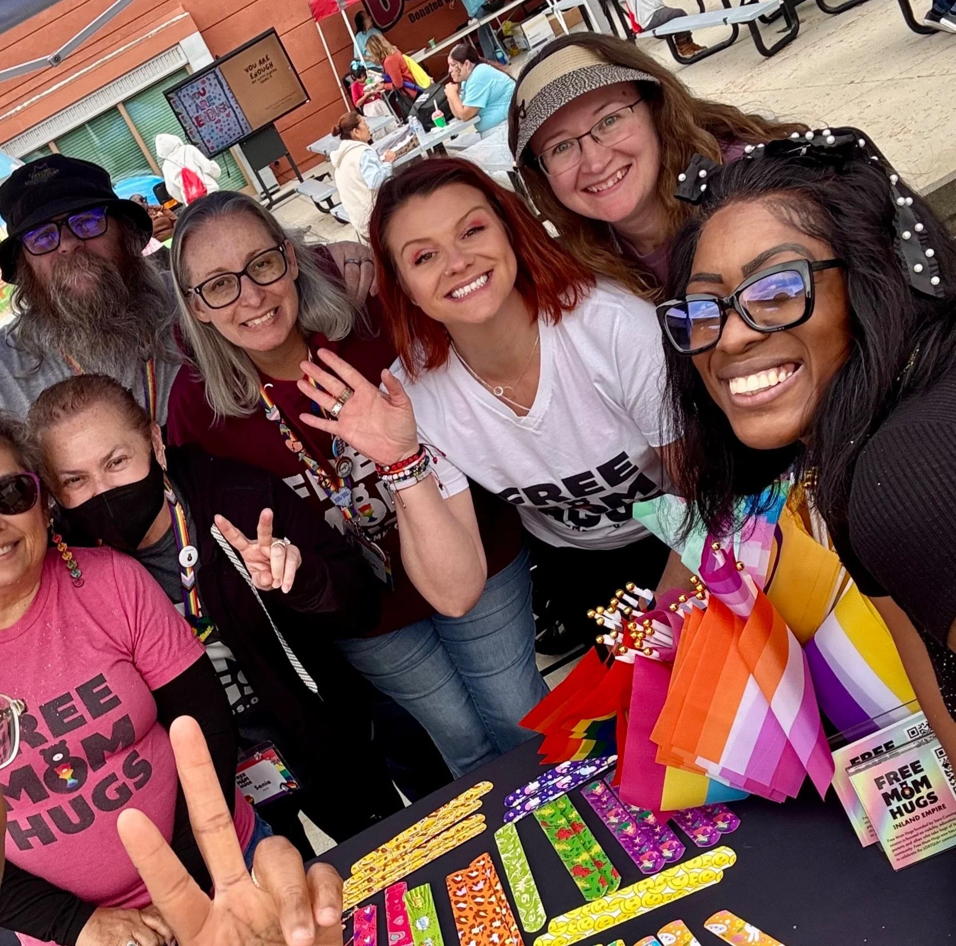 Group of diverse people smiling at a pride event, with a table of colorful pride-themed items in front of them.
