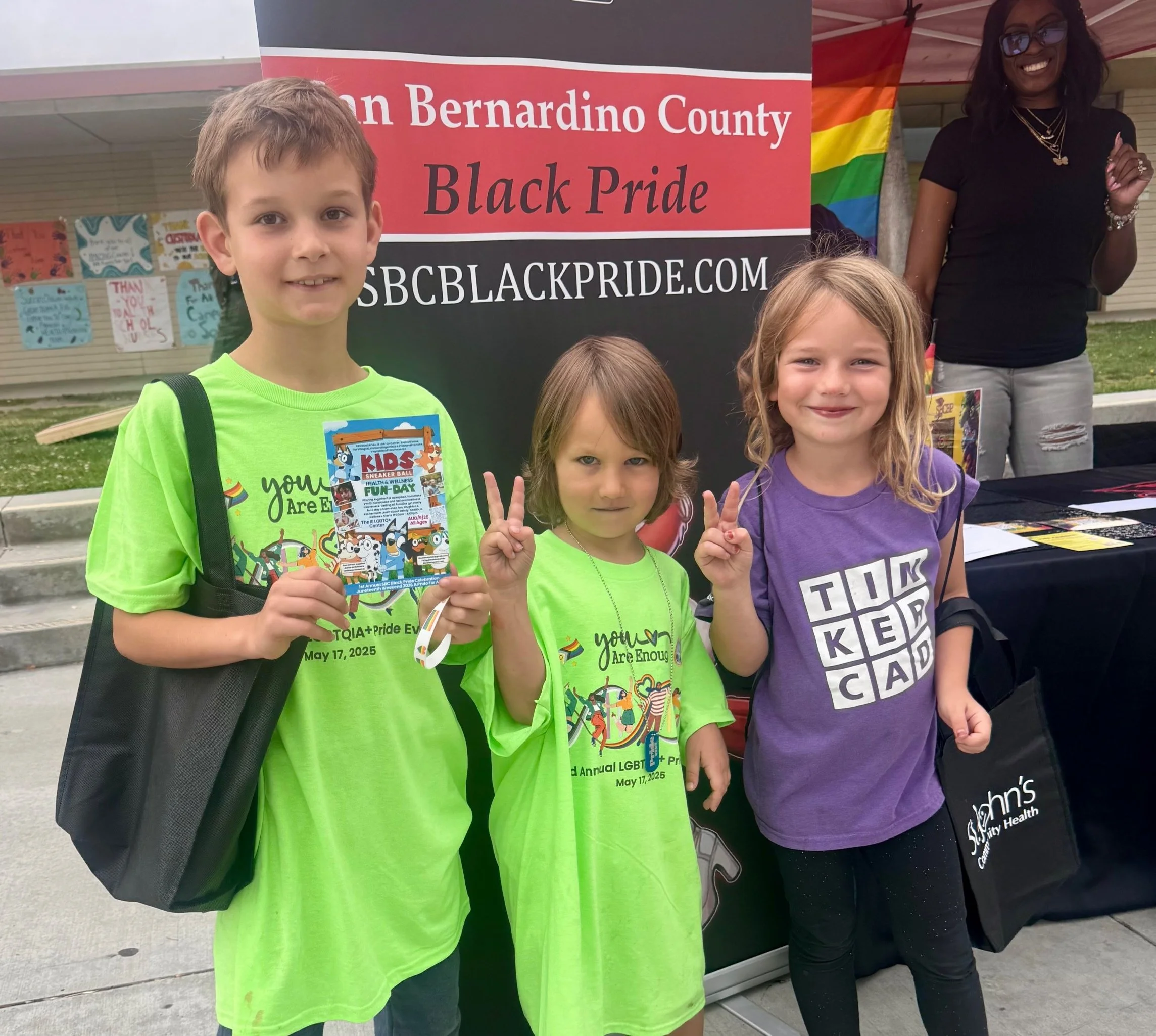 Three children standing in front of a Black Pride event banner, making peace signs. One boy and two girls are smiling, and the boy is holding a Kids sneaker ball event flyer. Two of the children are wearing bright green T-shirts, and the girl on the