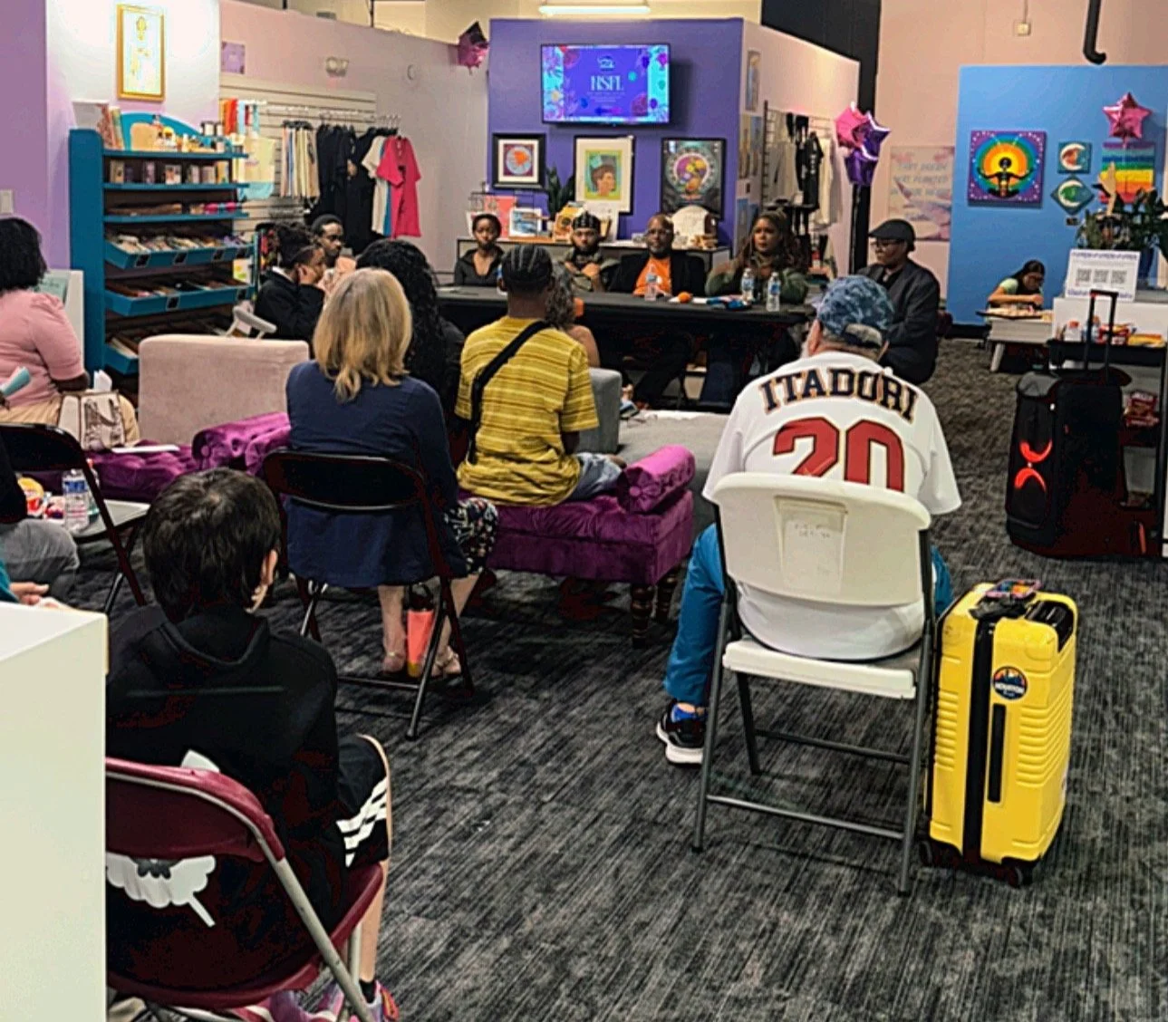 A diverse group of people sitting in a colorful indoor space, attending a panel discussion or meeting, with artwork and merchandise displayed on the walls.