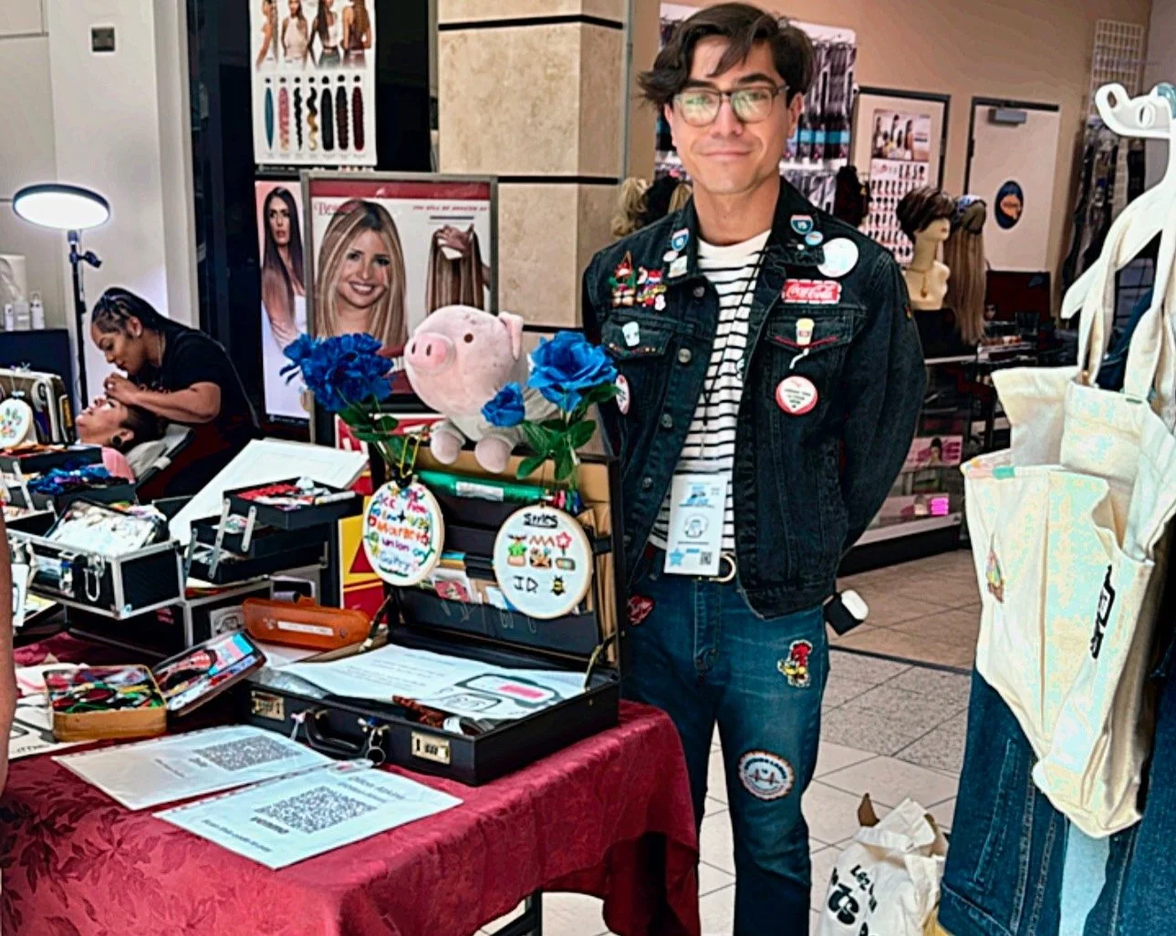 A person with glasses, wearing a denim jacket decorated with pins and a striped shirt, stands at a booth with various items including a plush pig, blue flowers, and embroidered circular patches. In the background, a woman is getting her hair styled,