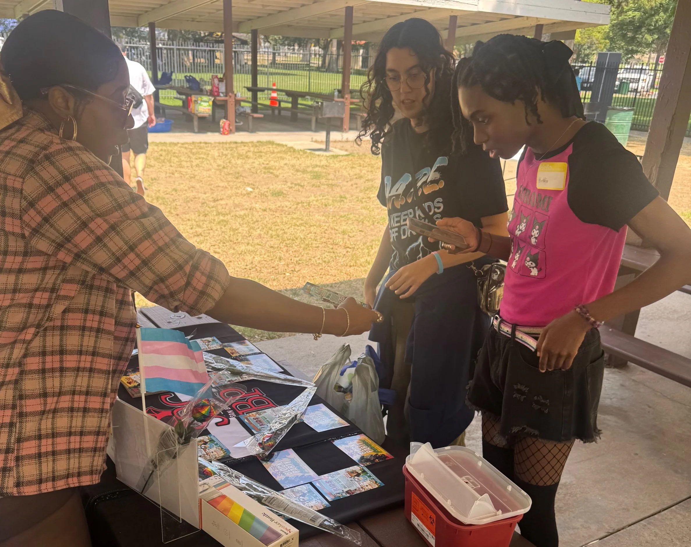 Three young women at a table under a pavilion outdoors, engaged in conversation; the woman on the left is handing her something, and the other two are looking at her. The table has various items, including pamphlets, a rainbow flag, and small toys. I