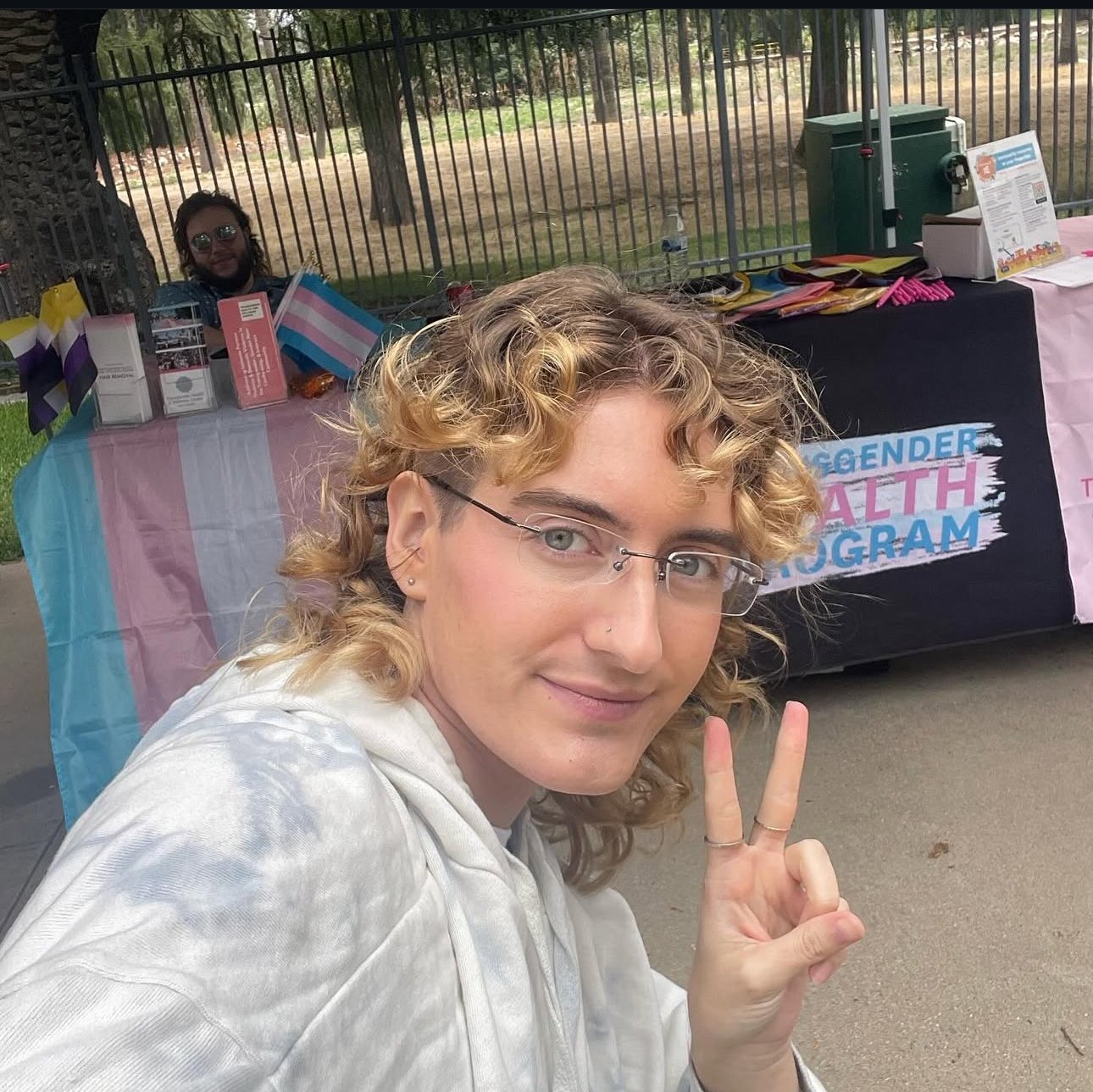 A person with curly blonde hair and glasses making a peace sign in front of a booth for a gender health program, with pride flags and informational materials visible.
