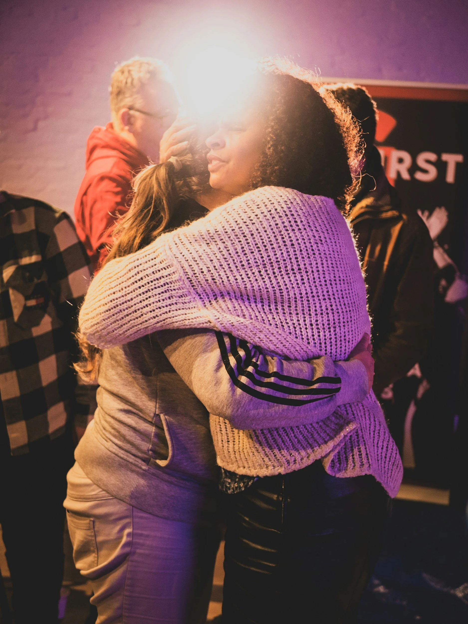 A woman with curly hair and a woman with brown hair hugging each other warmly at a social gathering or event.