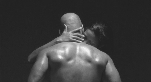 Two shirtless men embracing and kissing, one with his hand on the other's neck, against a dark background.
