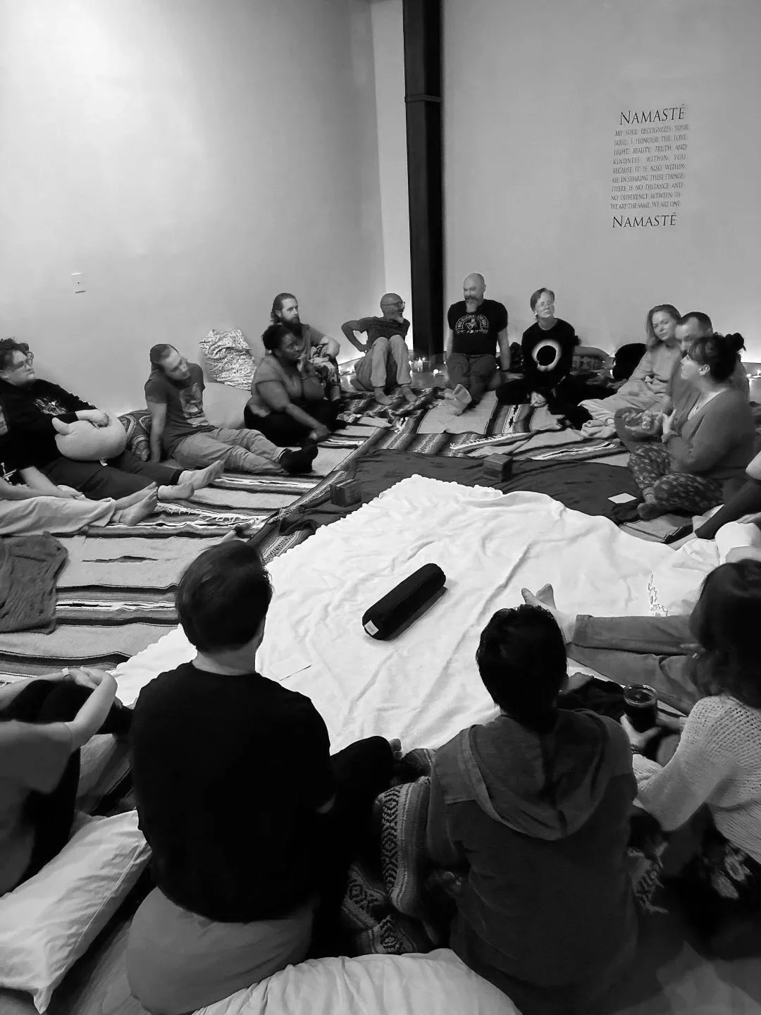 People sitting on the floor in a circle, engaging in a discussion or meditation, with a white cloth in the center, in a room with a Namaste quote on the wall.