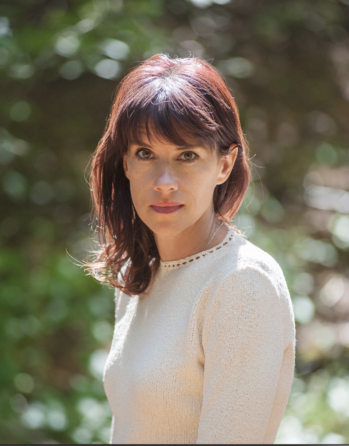 A woman with shoulder-length reddish-brown hair wearing a cream-colored sweater outdoors with a blurred green foliage background.