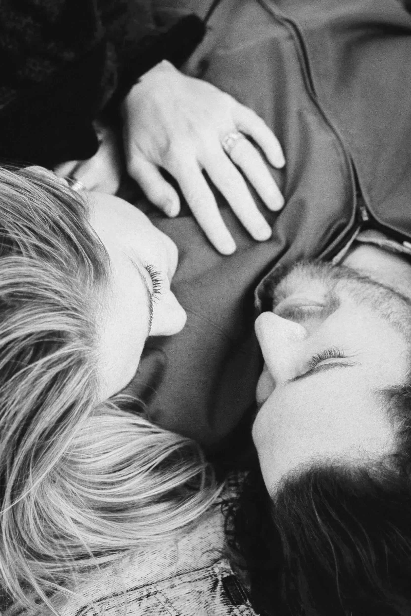 Two women with blonde hair, lying face to face with a dog between them, touching paws and noses, in a close-up black-and-white photograph.