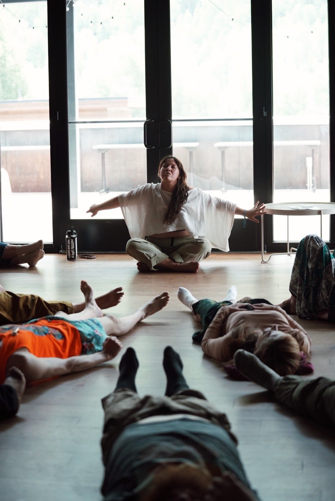 A woman sitting cross-legged on the floor with arms outstretched, leading a yoga or meditation class, surrounded by people lying down on the floor in a room with large glass doors and a balcony outside.