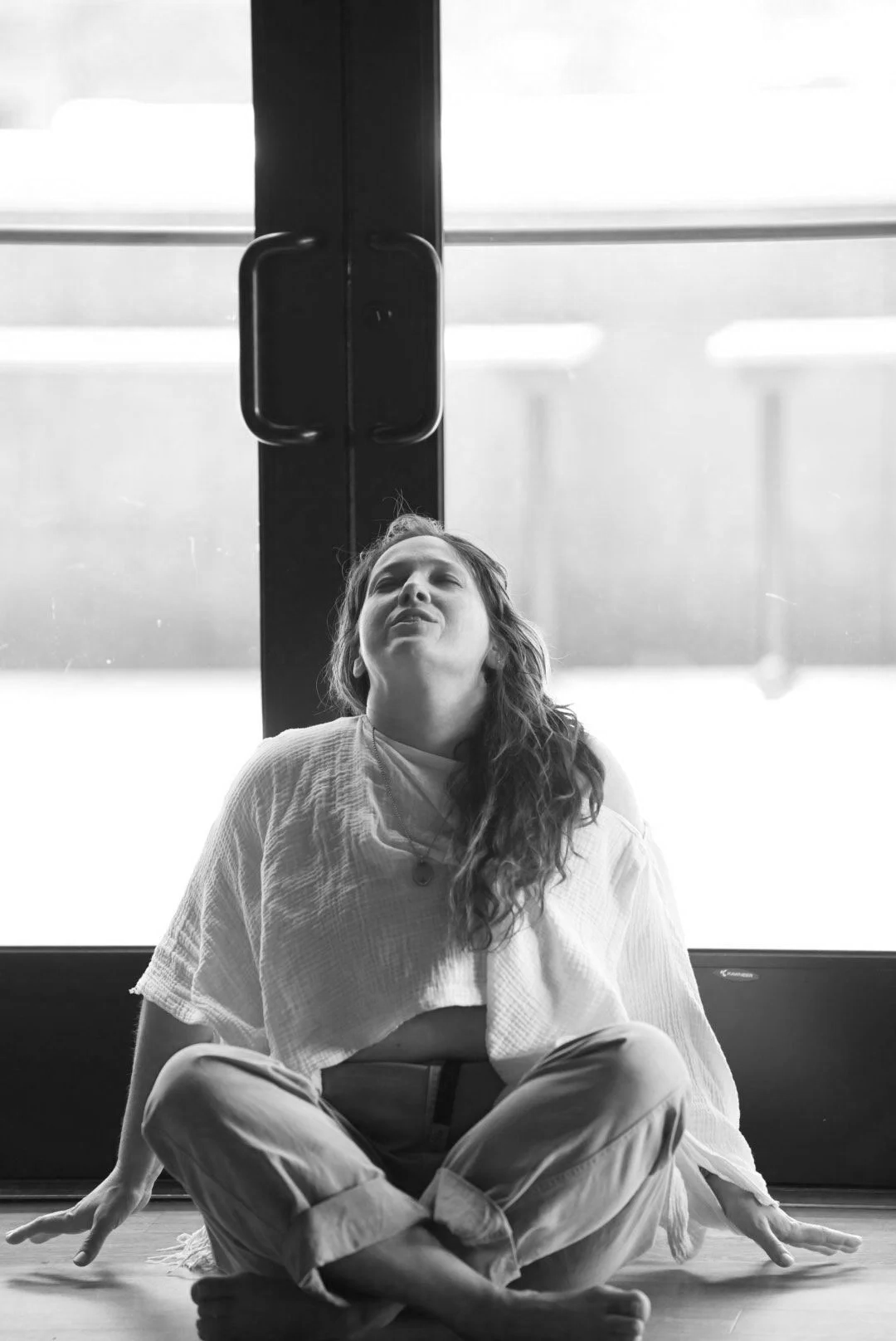 A woman sits cross-legged on the floor in front of a glass door, with her head tilted back and eyes closed, appearing to be meditating or relaxing.